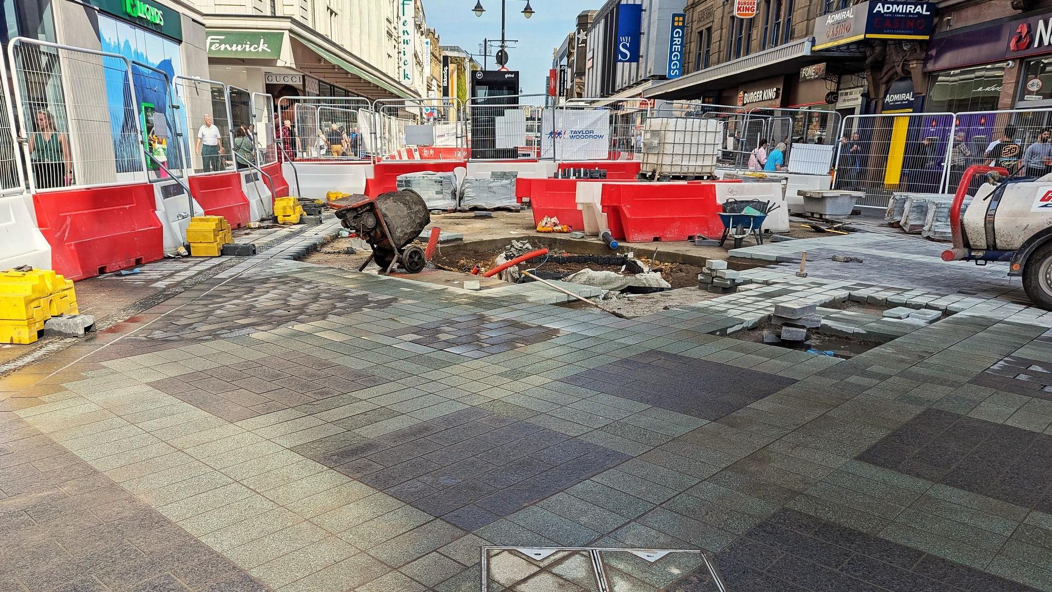 Bright red and white construction barriers surround a section of Northumberland Street. In it, half of the paving is laid down and forms grey diamond patterns. The other half is a work in progress where the street is still dug up and a cement mixer sits nearby.