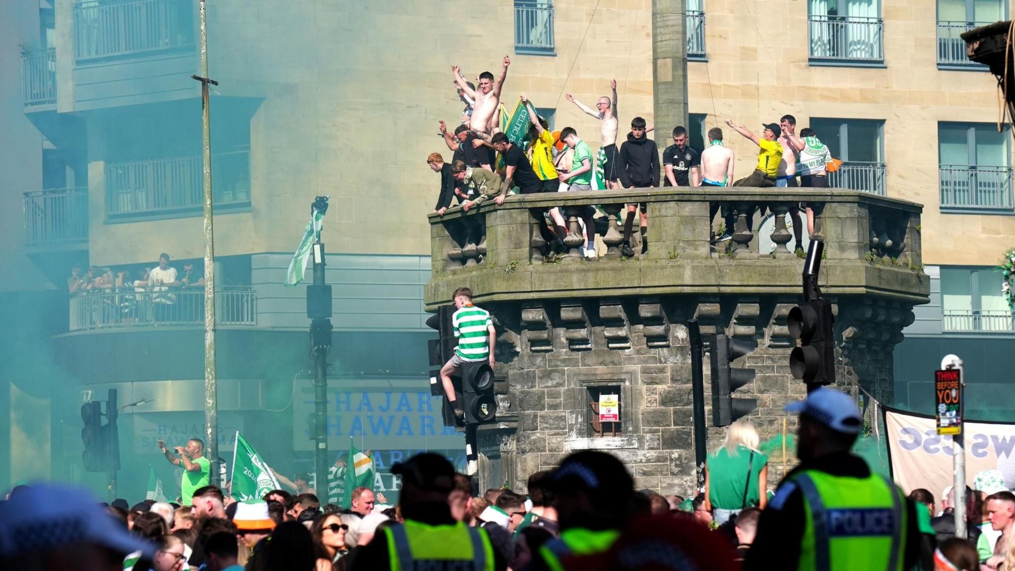 A large gathering of Celtic fans on the Trongate in Glasgow. A number of fans are on top of the Mercat Cross and on traffic lights. Smoke fills the air. The fans are wearing Celtic strips and waving Celtic flags