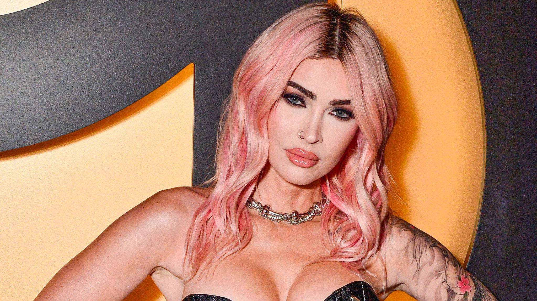 Megan Fox staring at a camera. Wearing a gold necklace, with pink hair and tattoos on her left arm. She's stood in front of an orange and black background. 