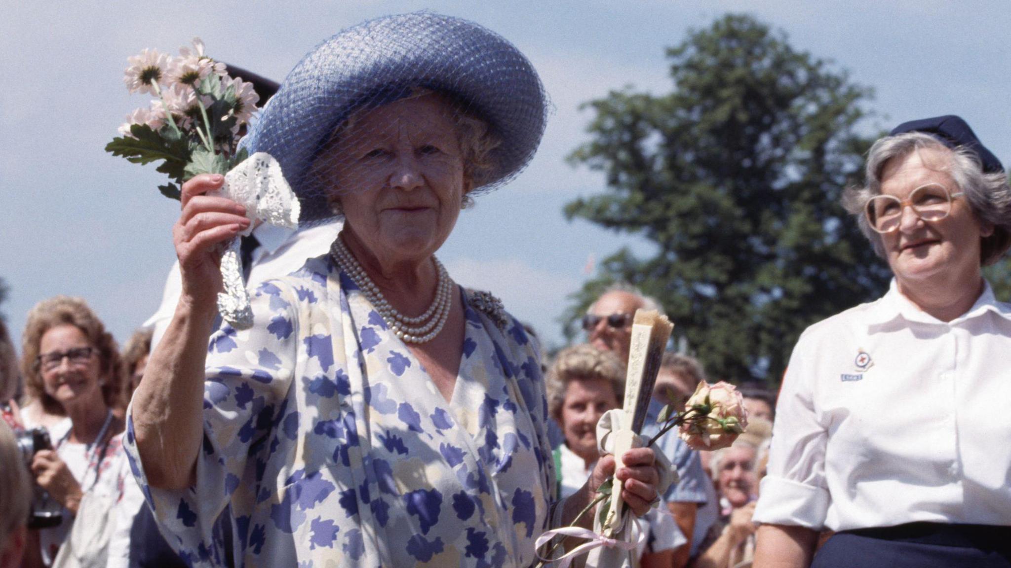 The late Queen Mother holds a posy of daisy-type chrysanthemum flowers.
They are white and are held together by white lace material. Several woman stand behind the Queen Mother who also holds a fan and a single rose in the other hand. She is wearing a cornflower blue hat with matching netting and a white and cornflower-blue dress with a triple string of pearls around her neck.