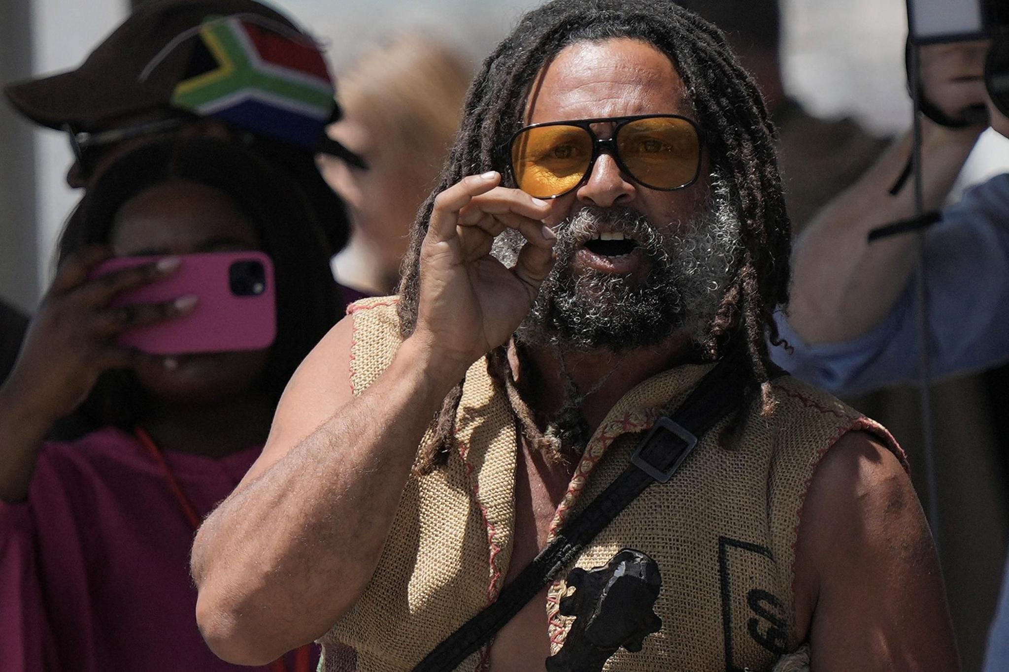 A protester with dreads, a beard and wearing orange lensed sunglasses shouts at Kalk Bay Harbour in Cape Town, South Africa - Thursday 7 November 2024 