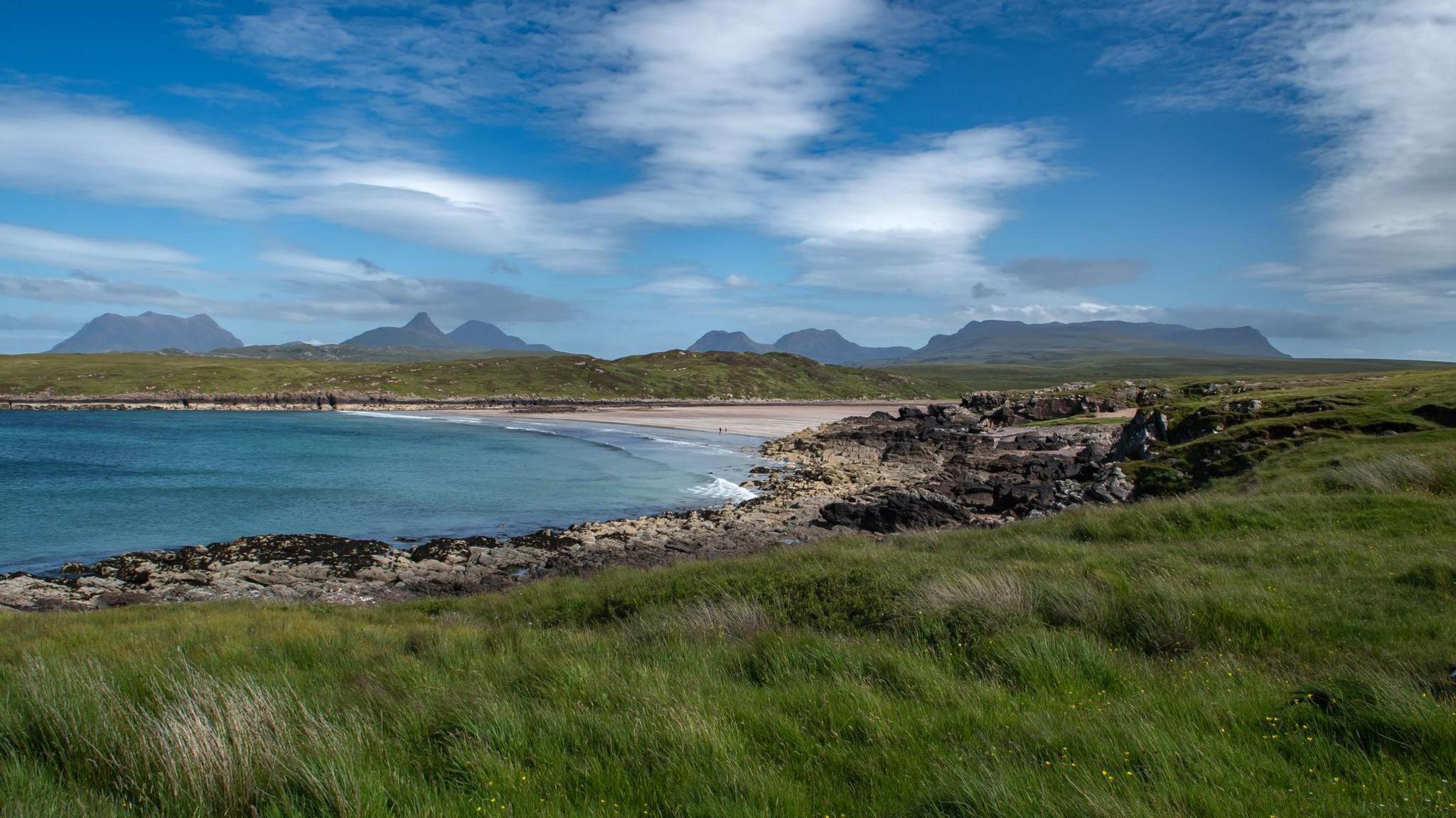 Clear blue waters of the sea and a beach with sand and rocks. In the distance are mountains under a blue sky with clouds.