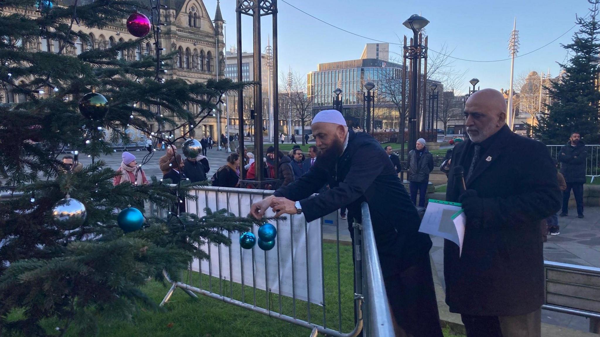 A man in a white skullcap with a beard attaching a blue shiny bauble to a branch of a fir tree with another man in a dark coat and black shirt and tie watching on.   