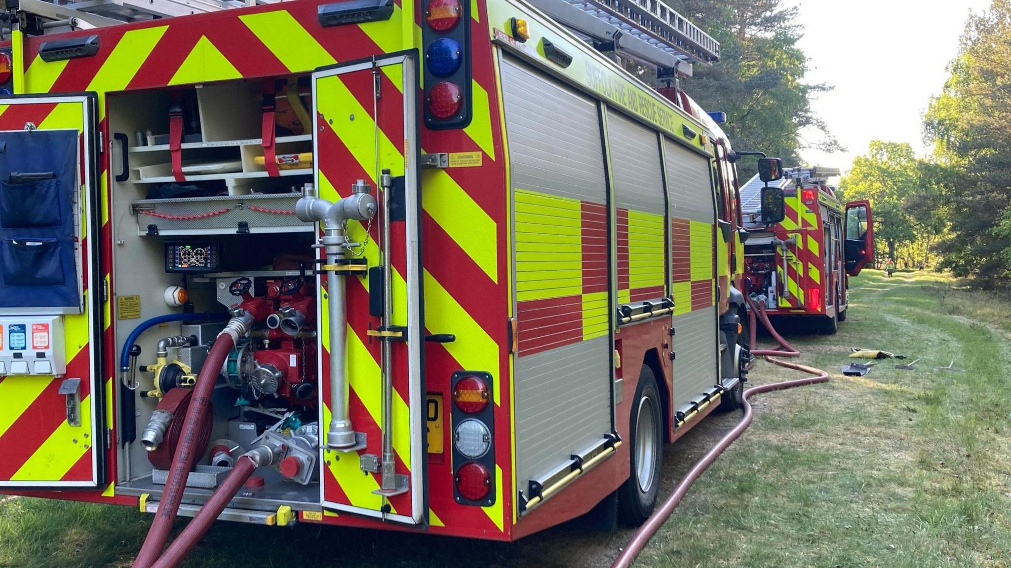 Rear view of two fire engines that have hoses coming out of the back. They are stood on green grass in the middle of a forest, with trees surrounding them. 