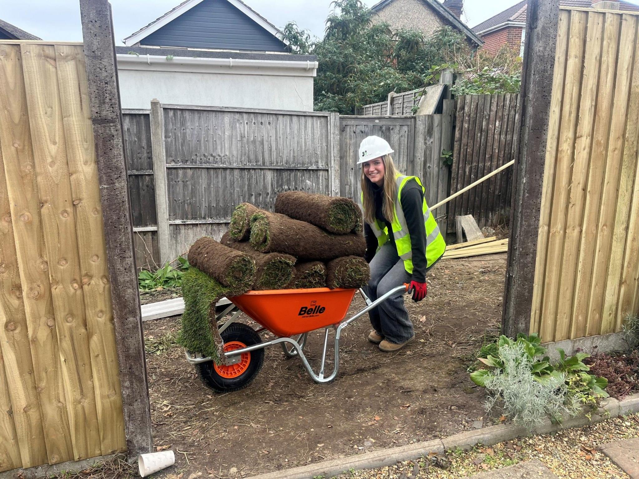 A woman wheeling an orange wheelbarrow full of rolls of turf. She is standing between two fence panels smiling. 