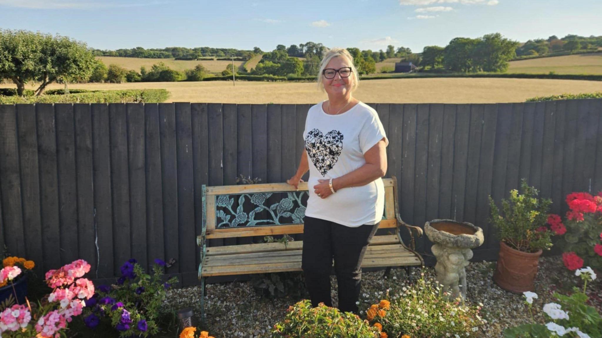 A woman with blonde hair in a ponytail and black glasses is smiling. She is standing in front of a wooden bench, which is in front of a fence - beyond which are an expanse of farmland fields and trees. In the garden in front of here are flowers and plants, as well as a bird bath