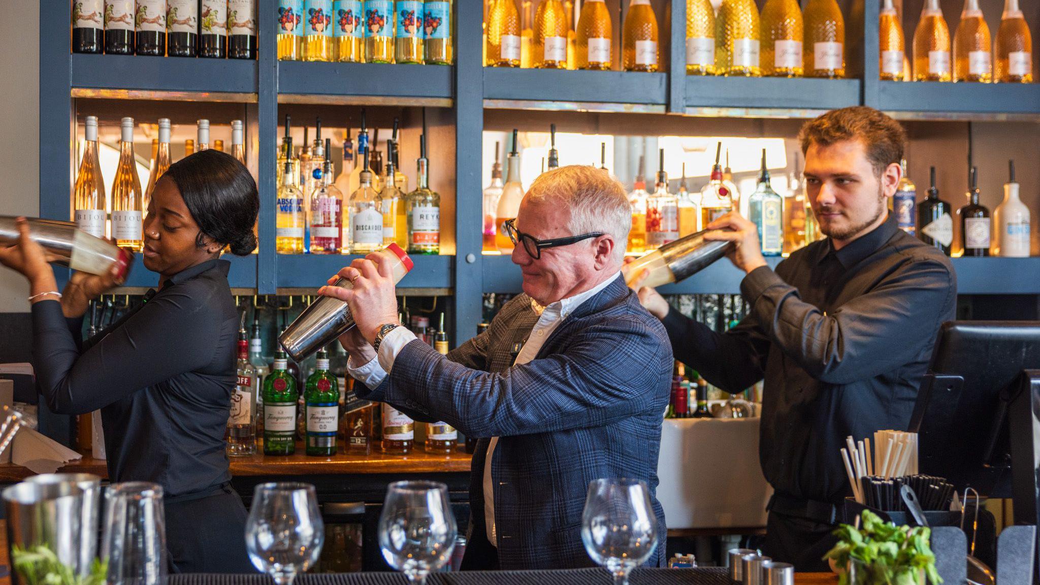 Two men and a woman stand in a line behind a bar, all shaking cocktail mixers. They are in front of blue shelving containing rows of bottles.