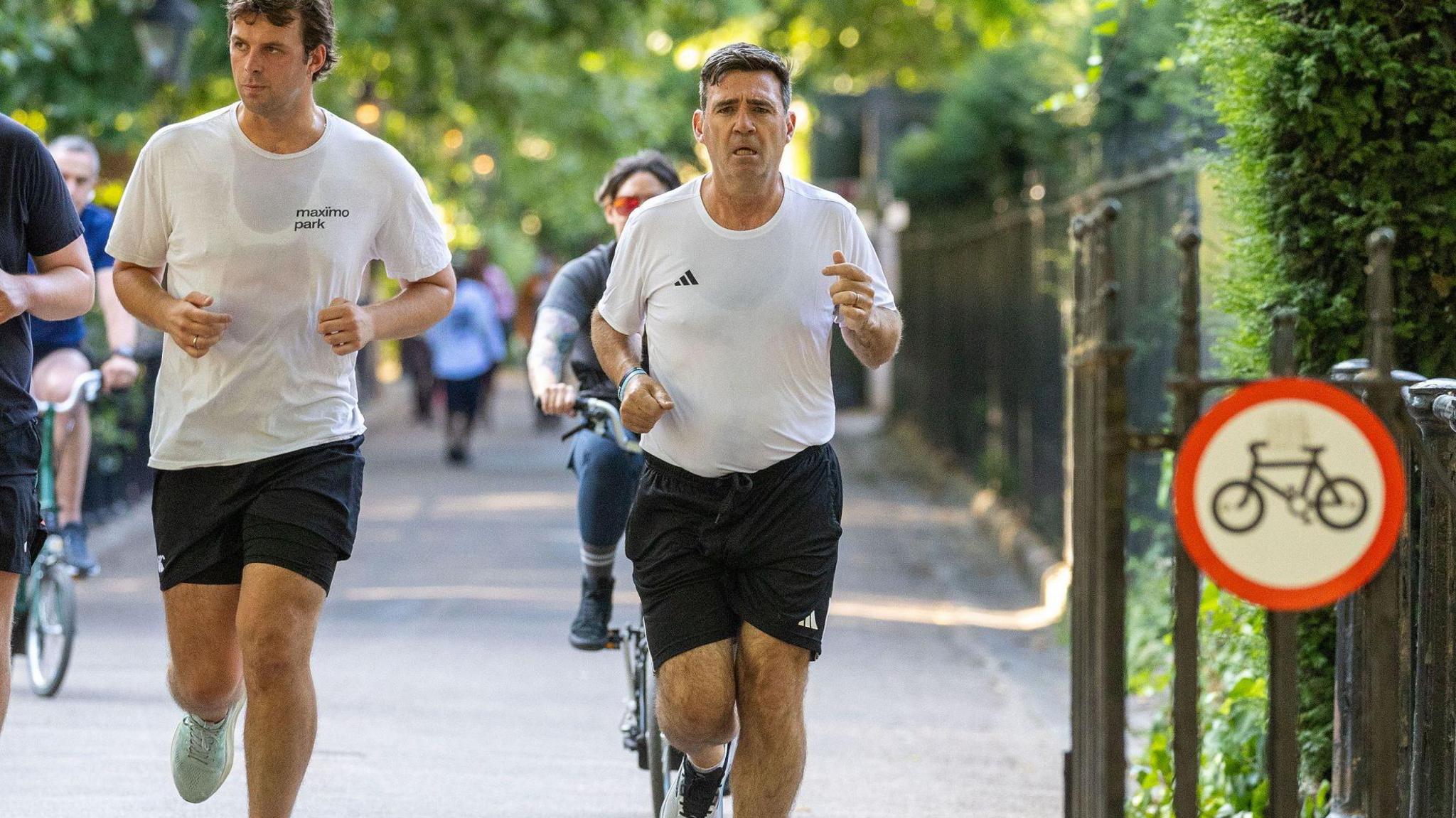 Andy Burnham, wearing a white Adidas top and black running shorts, running along a pavement in Green Park