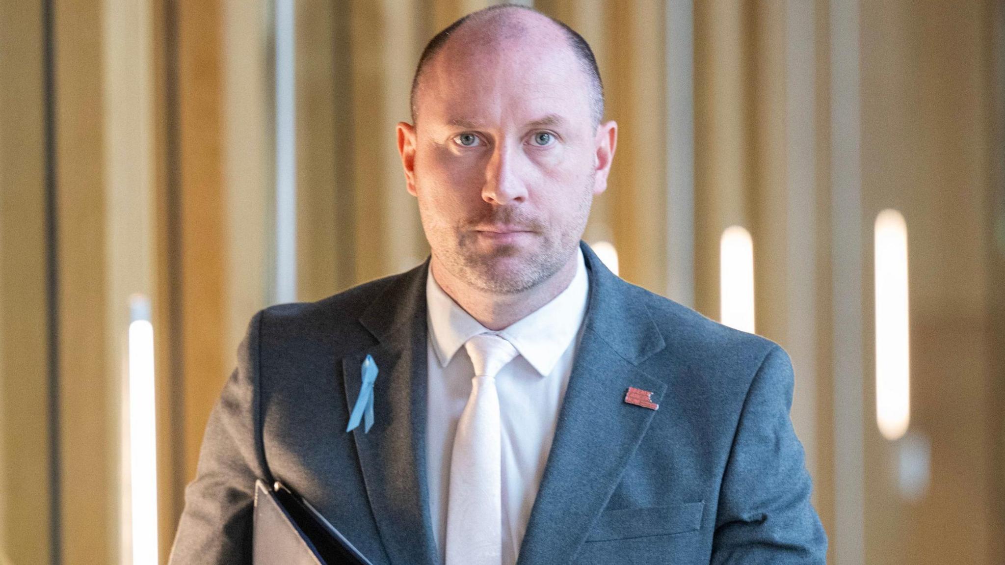 Neil Gray, a bald man with his hair shaved at the sides, walks along a well-lit corridor. He is wearing a grey suit, peach shirt and matching tie. He has a serious expression on his face and his staring directly at the camera. 