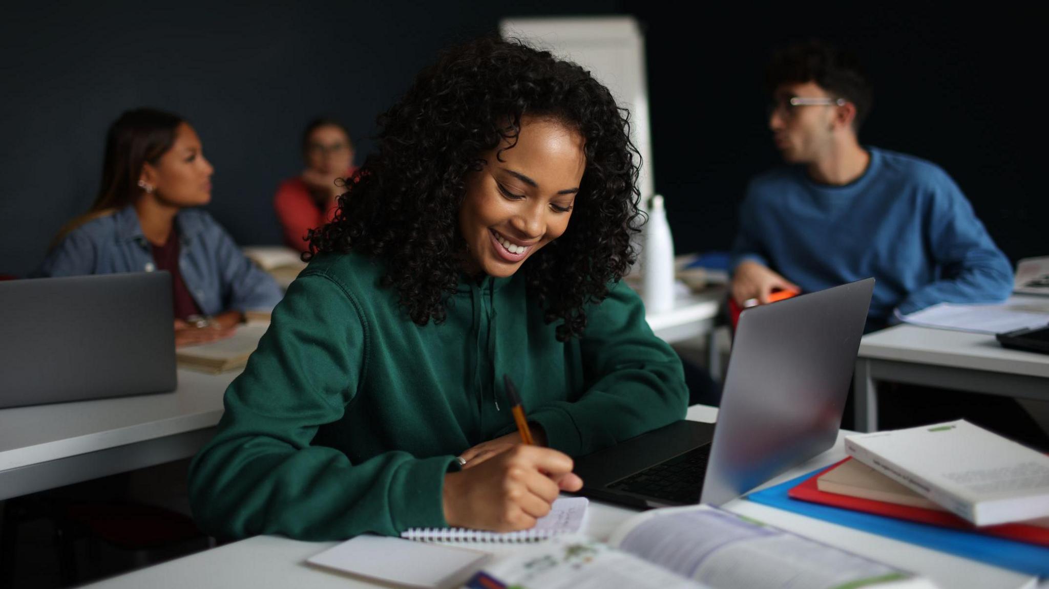 The picture shows a young woman writing in a notebook during a university lecture. She sits in a classroom with other students and has a laptop in front of her. 