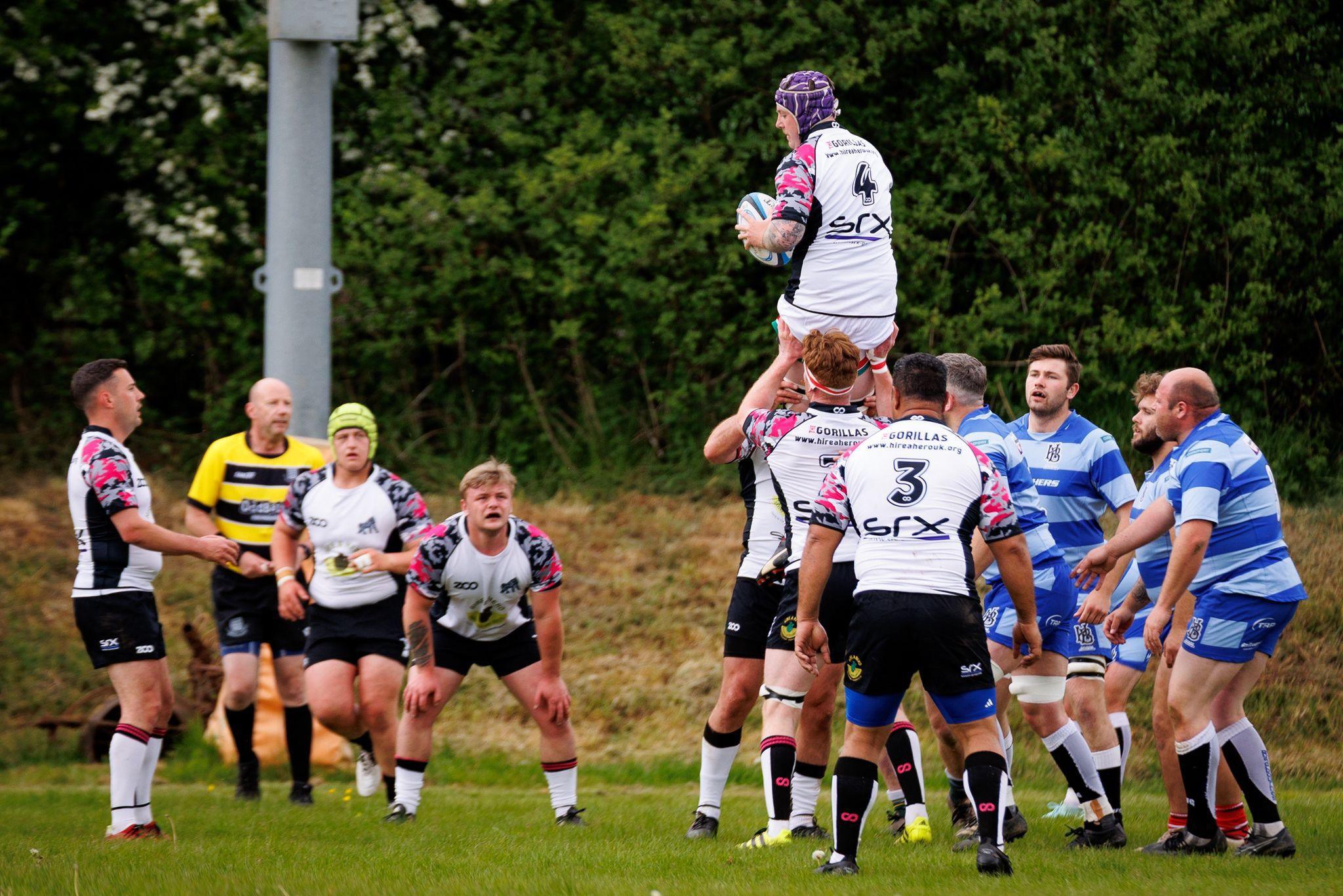 A match between the Hospice Barbarians and The Gorillas RFC. Players from both teams are seen in their different kits. One man is on another man's shoulders. They are on a rugby pitch with a post and trees in the background.