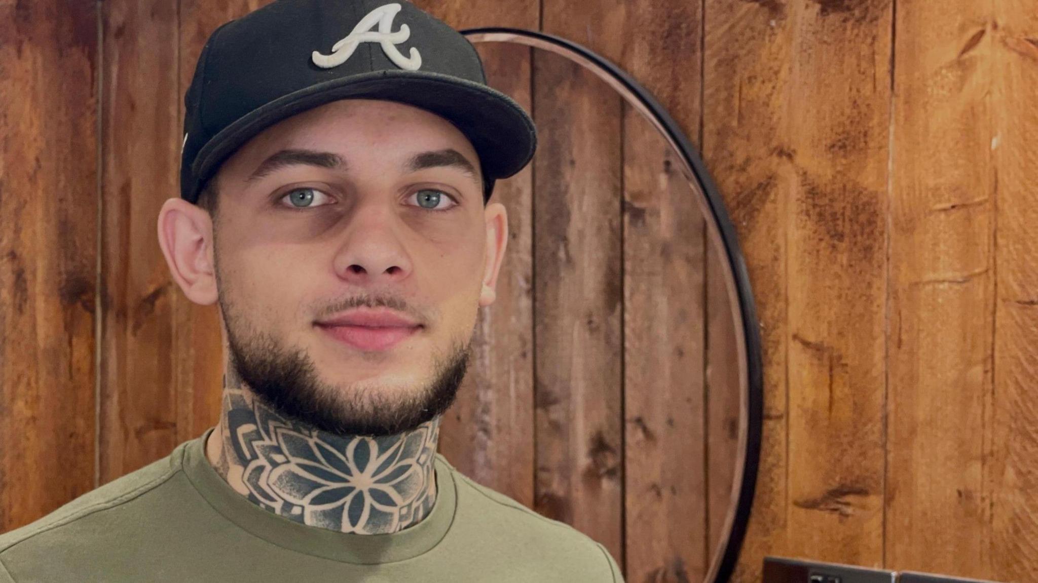 A man with a short beard wearing a baseball cap looks directly at the camera. He is standing in a barber shop and has a distinctive flower-based neck tattoo.