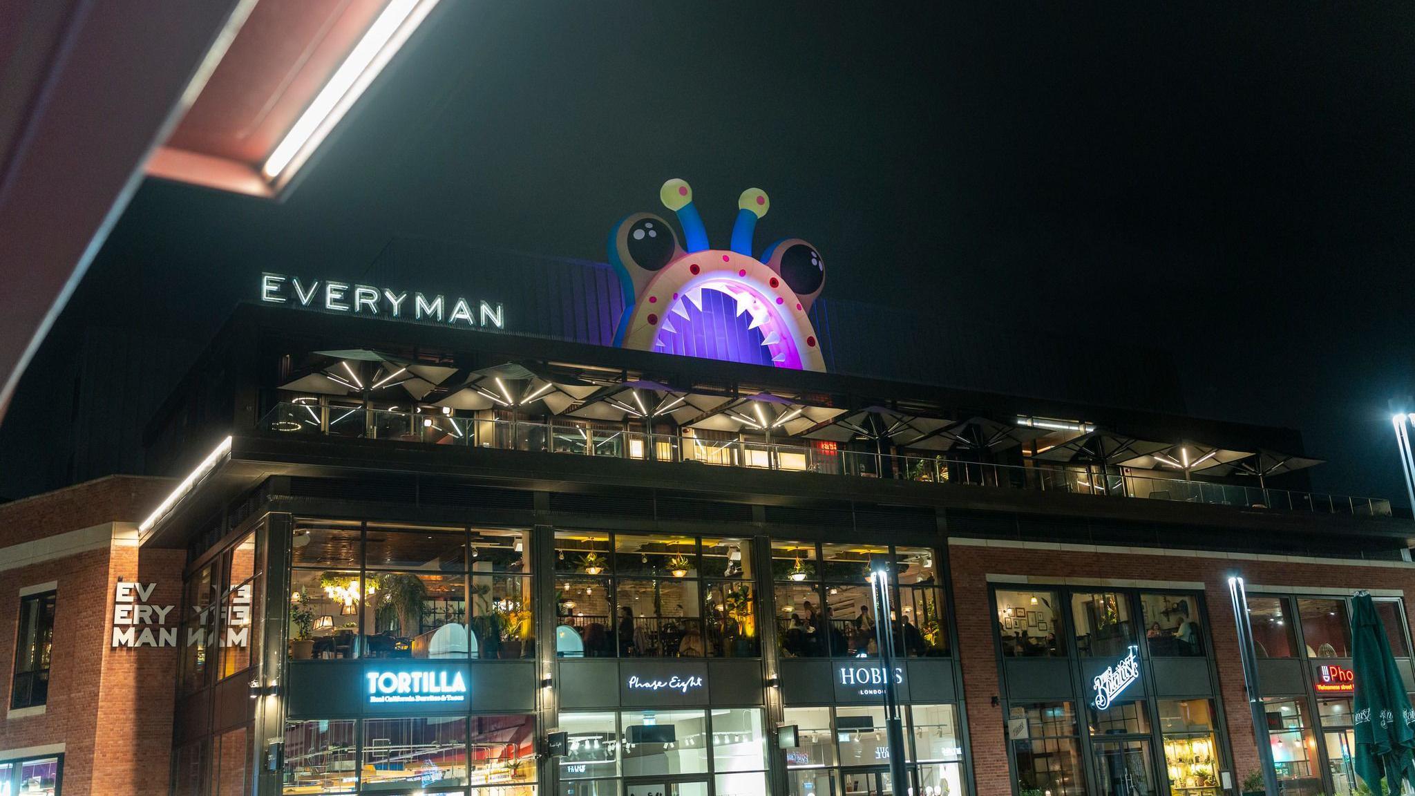 A white arch with white teeth with black eyes and blue and green antennae's makes up an inflatable monster which is lit-up and on top of Everyman Cinema. It is night time and shops below include Tortilla, Hobbs and the Botanist.