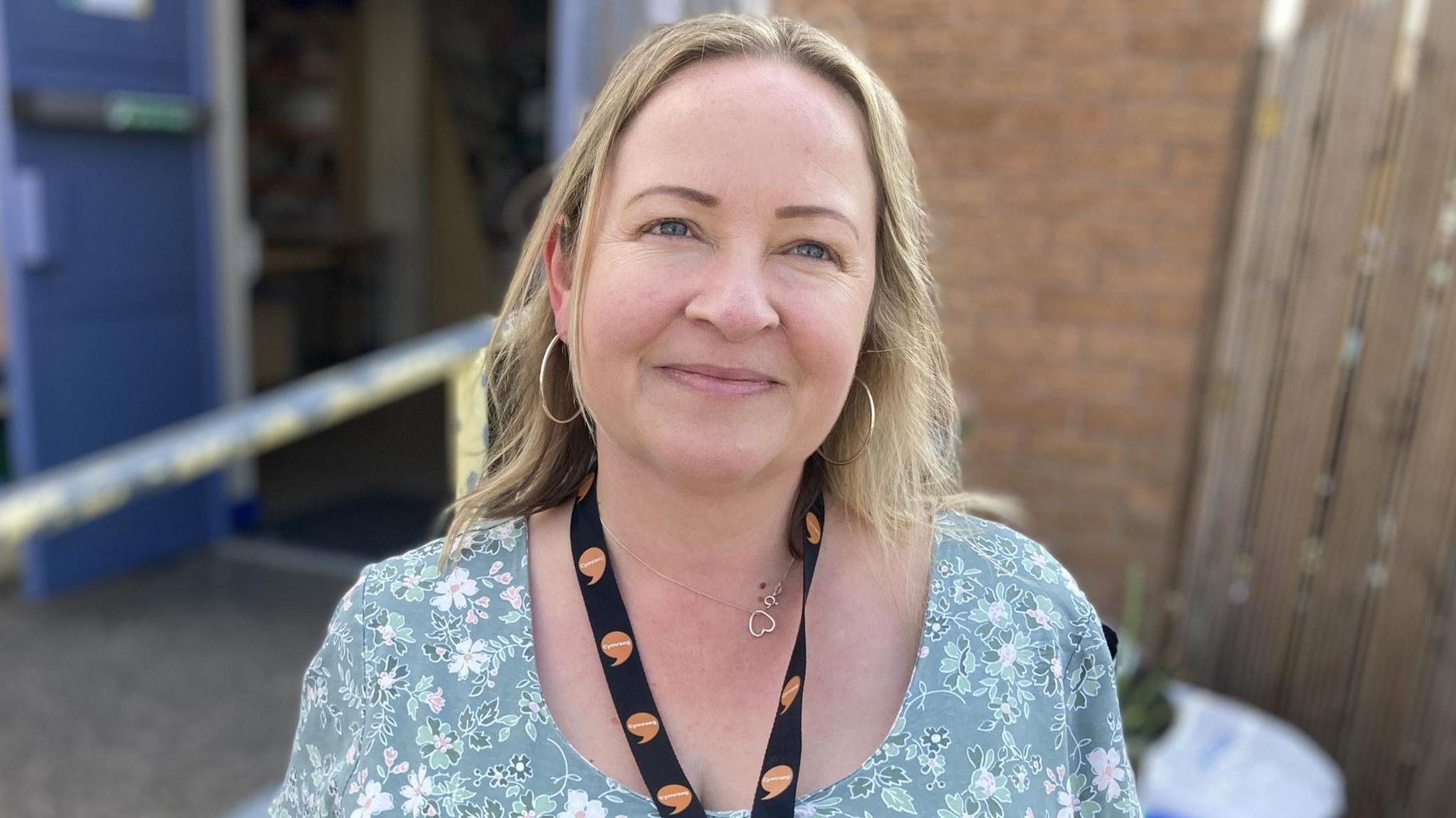 Lynne Thomas yn edrych at y camera mewn ffrog flodeuog, gyda adeilad gymunedol tu ol iddi