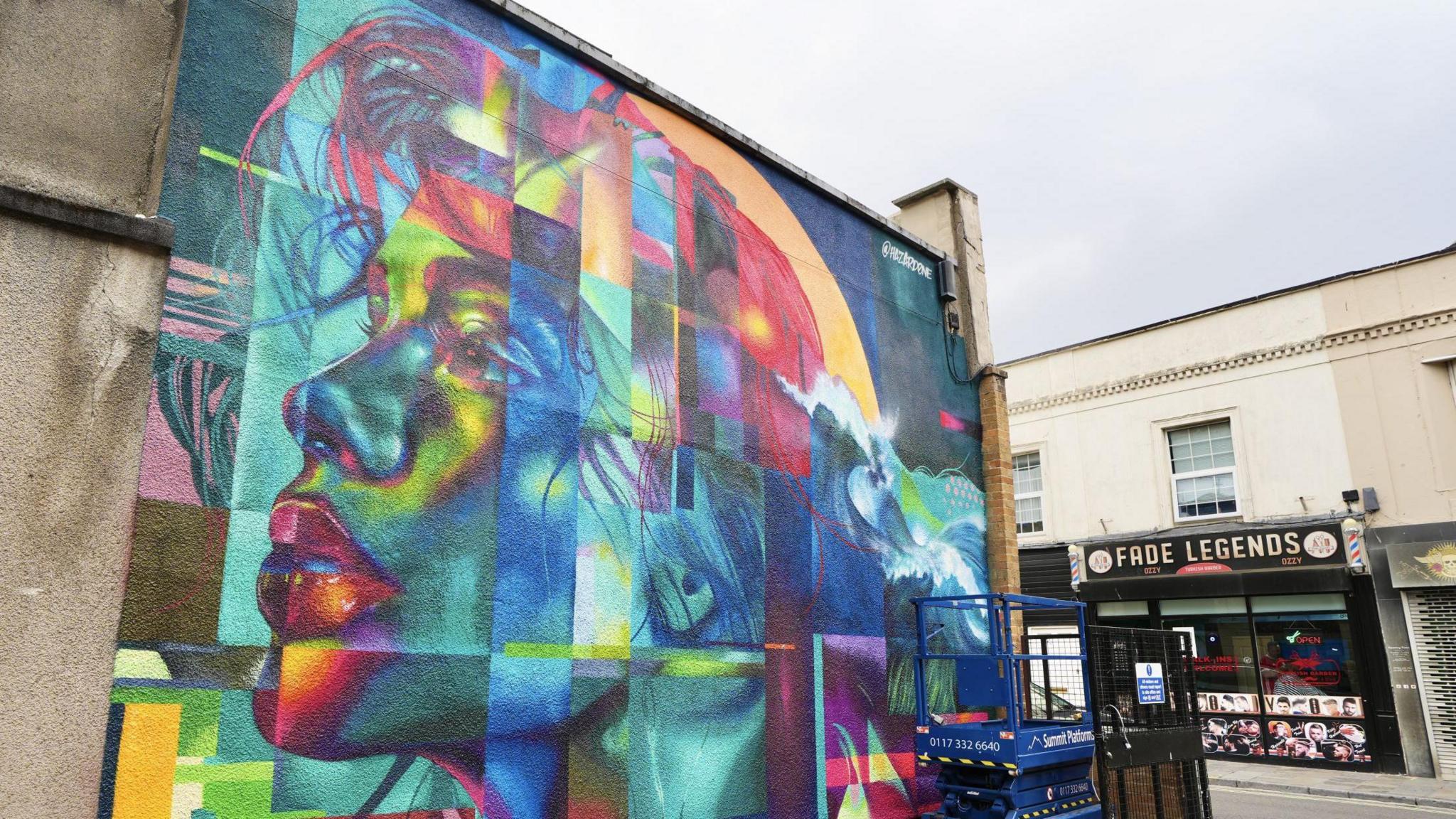 A large multicoloured mural of a woman's side-on profile on a wall in Weston-super-Mare. The mural is made up of different blocks merged together. In the background a barber shop with "Fade Legends" on a sign above the door is visible
