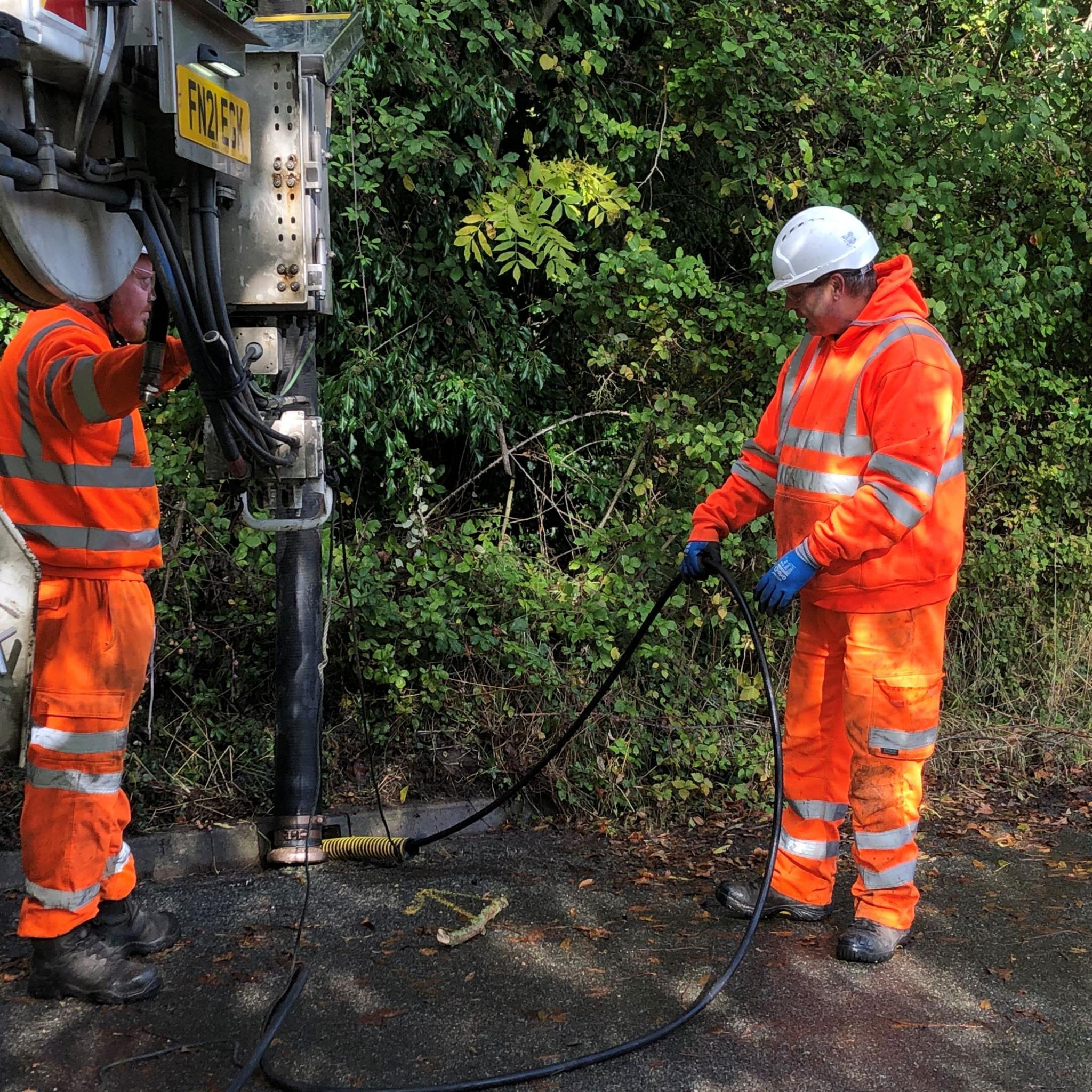 Two men in orange protective suits and hard hats clear out a roadside drain using specialist equipment. The path borders extensive green-leaved bushes. 