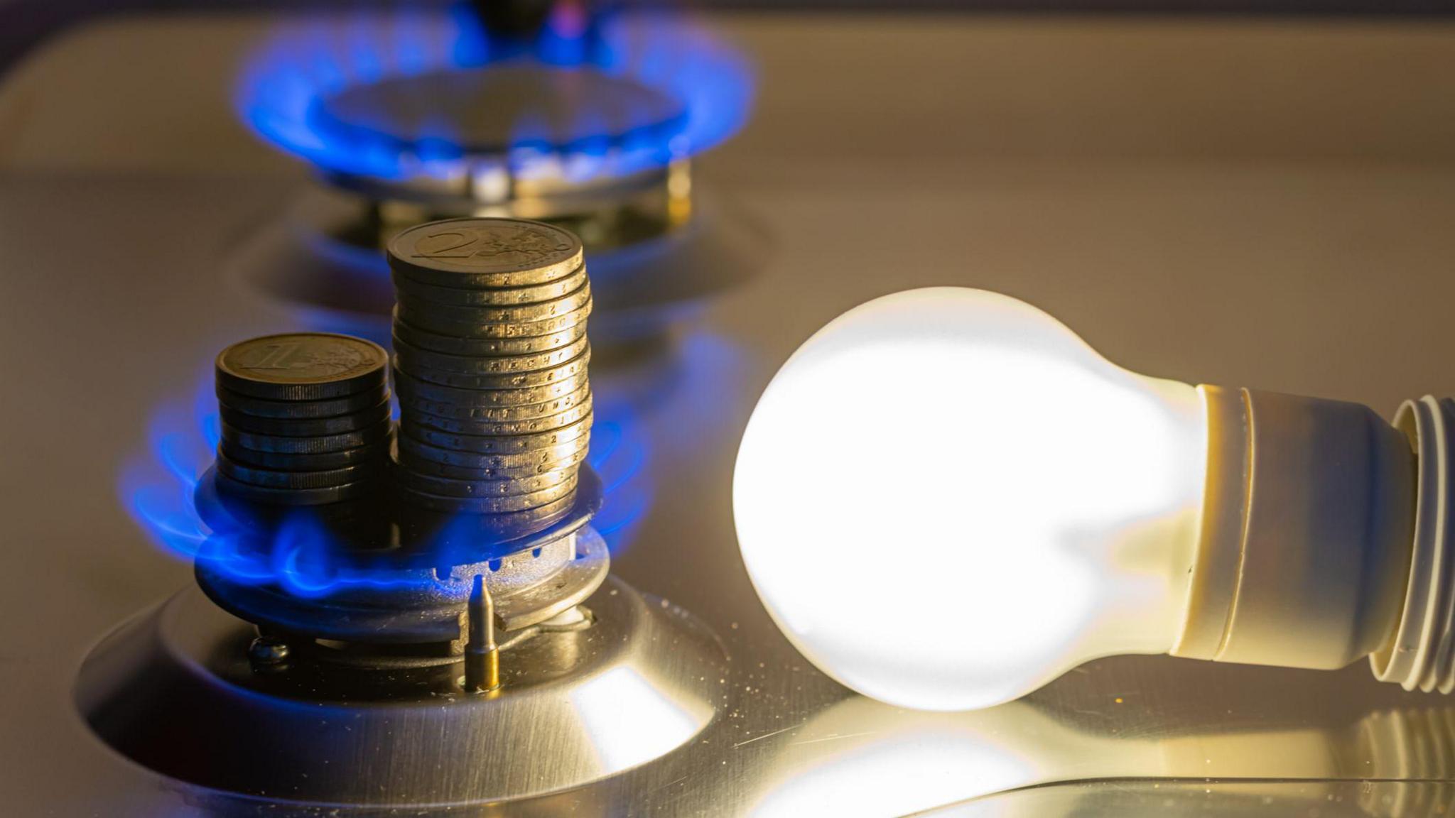 A lightbulb and a gas hob are lit next to each other. There are stacks of coins on the gas hob.