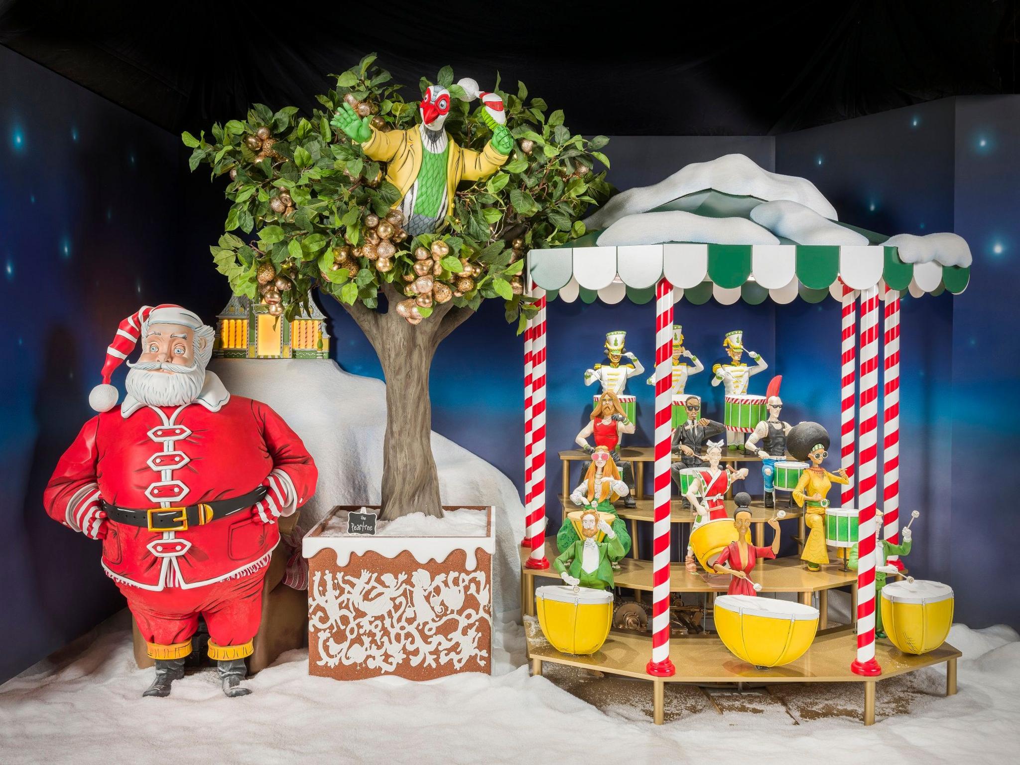 Santa in a red costume is standing next to a pear tree, which is next to a stand filled with figures banging on drums which are yellow and green.