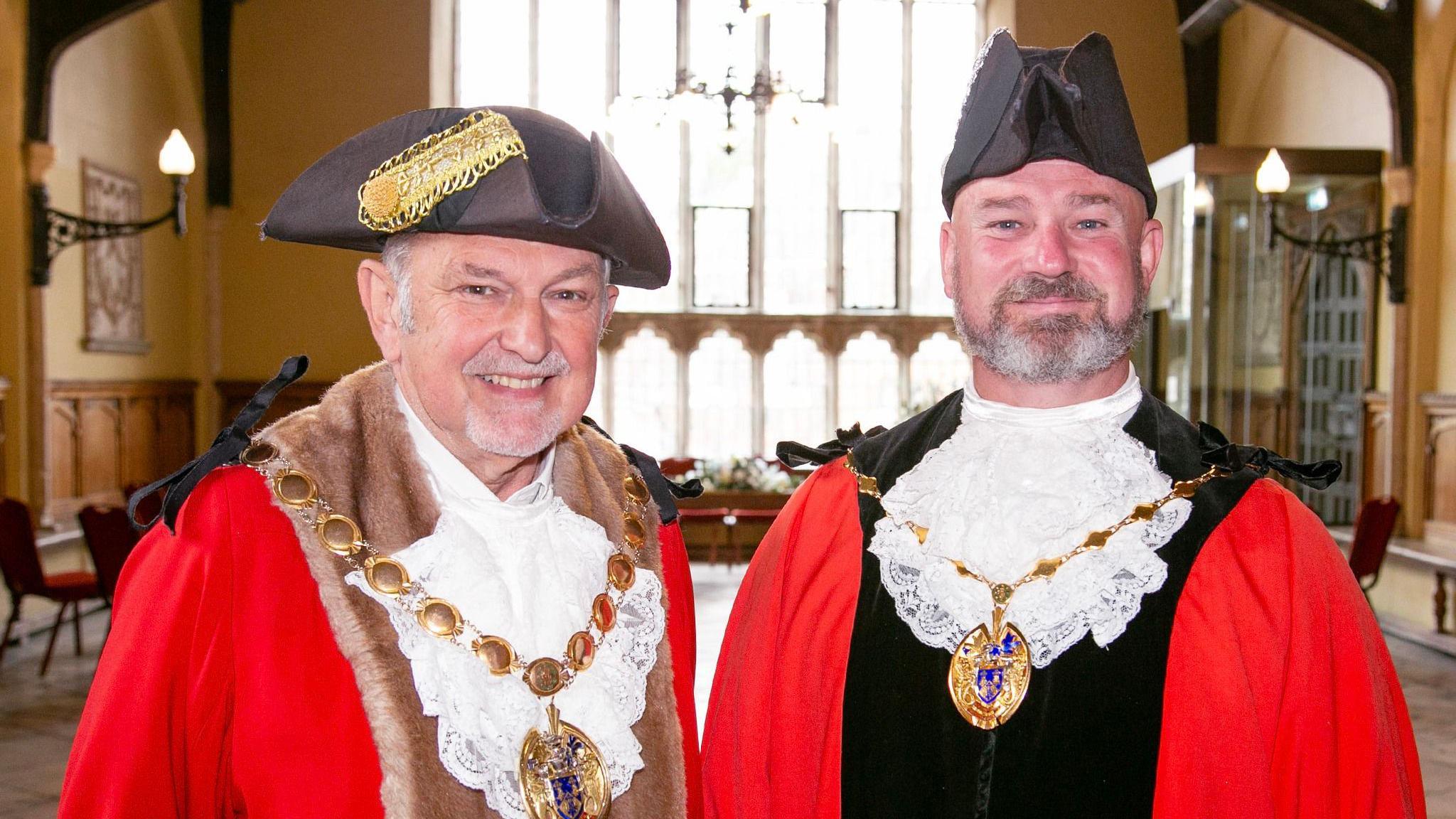 We can see two men dressed in mayoral outfits. They are both wearing red robes, tricorn hats and gold chains