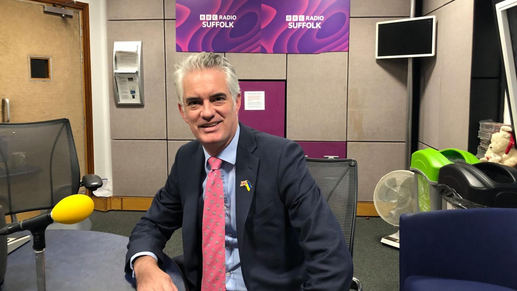 James Cartlidge MP wearing a dark blue jacket, light blue shirt and pink tie sitting at a table with a yellow microphone in the BBC Radio Suffolk studios.