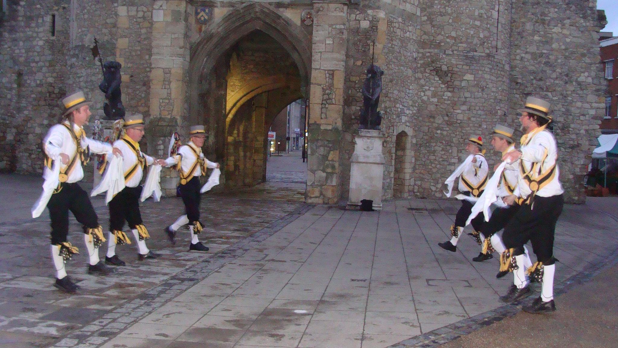 Six Morris men from King John's Morris Men wearing top hats white shirts, black trousers and white socks pulled up to the knee. They have yellow and black tassels and accessories. They are dancing in front of the bargate in Southampton at dawn