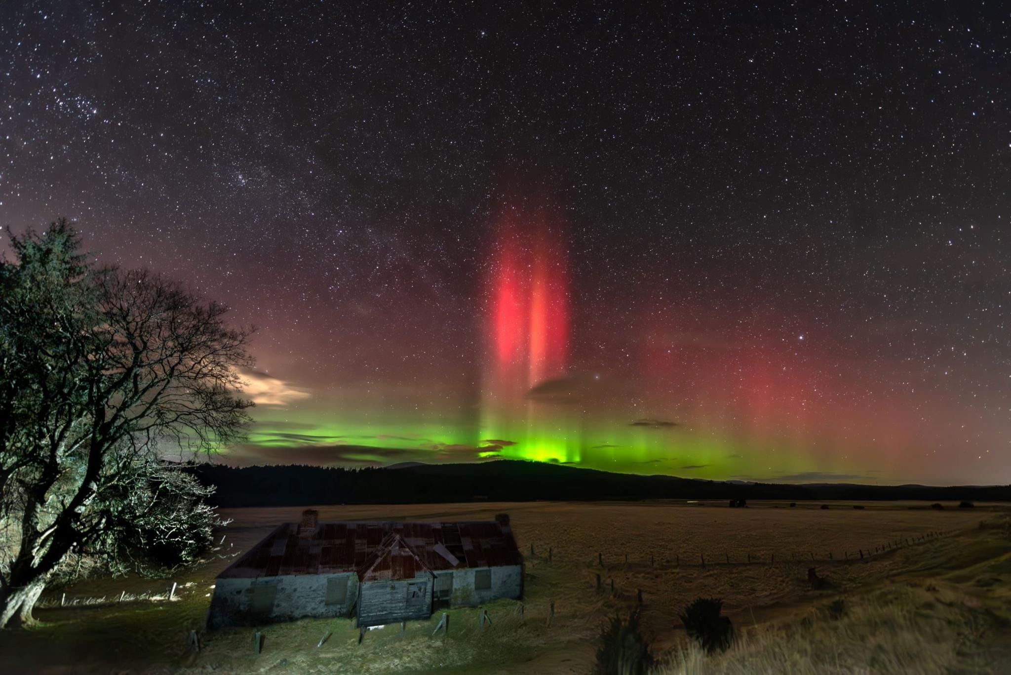 Red and green coloured northern lights in a starry sky with an empty field and small stone farm building on the left with a large tree in the front of the house