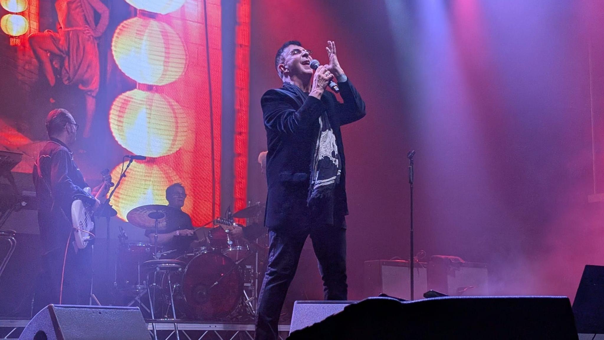 A man stands on stage and sings into a microphone. The stage has red and pink lighting and a band. The man has short dark hair and is wearing a black jacket.