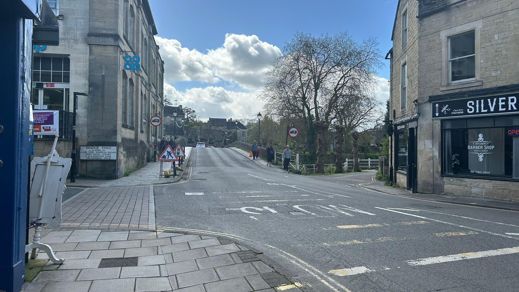 A view of Silver Street in Bradford-on-Avon, with people walking over bridge in background and line of sandy-coloured buildings near road.