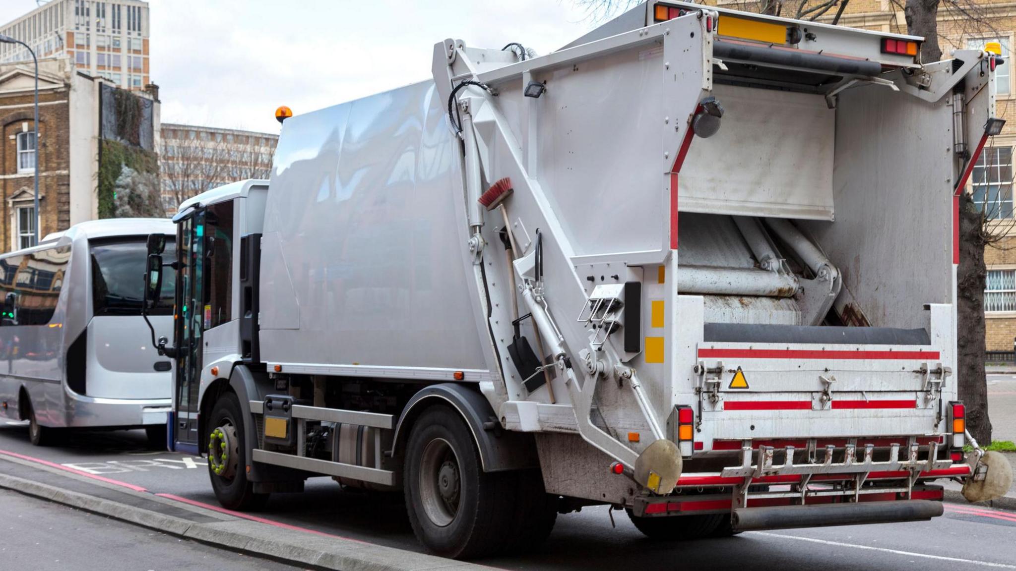 A white bin lorry drives down a street. The lorry is mostly white, with dark wheels and red and yellow safety markings on the back. A white minibus with dark windows is driving ahead of it.