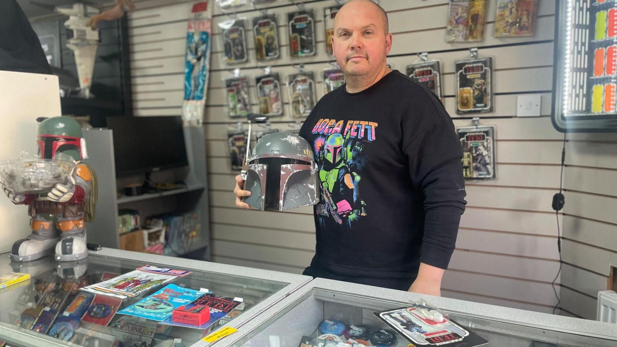 A bald  man in a black sweater stands behind the counter at a shop. The glass cabinet below him contains brightly coloured toys.