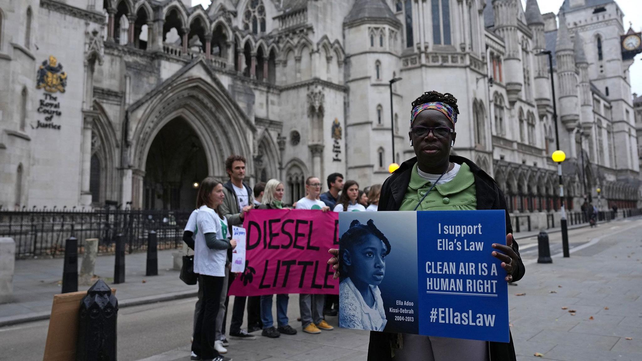 Campaigner Rosamund Adoo-Kissi-Debrah joins protesters outside the Royal Courts of Justice in central London - the court is visible behind her, and she is holding up a sign reading 'I support Ella's Law' and 'Clean air is a human right' with a picture of nine-year-old Ella Adoo-Kissi-Debrah