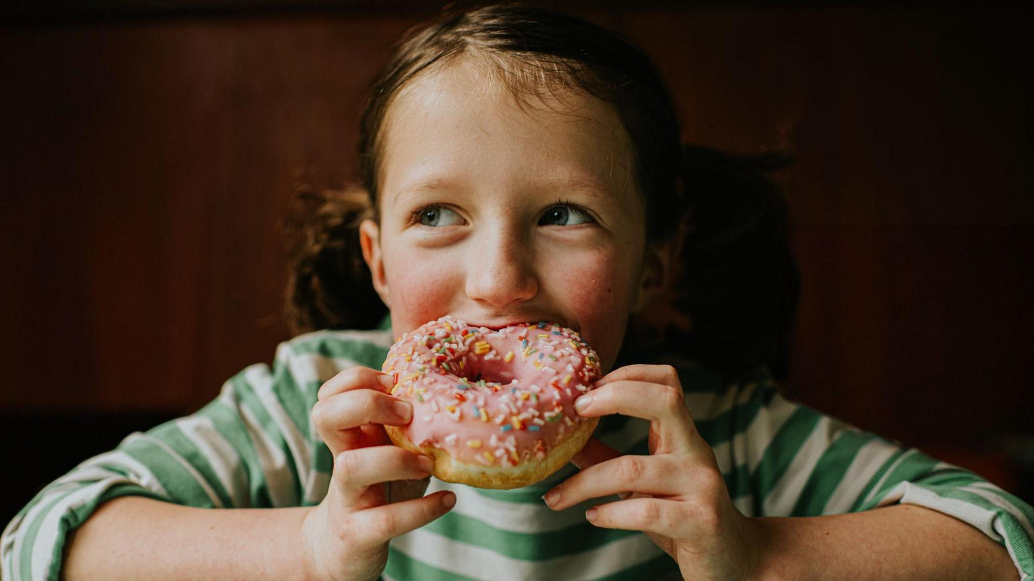 A child with brown pigtails and wear a green and white stripped top biting into a pink sprinkled glazed doughnut. 