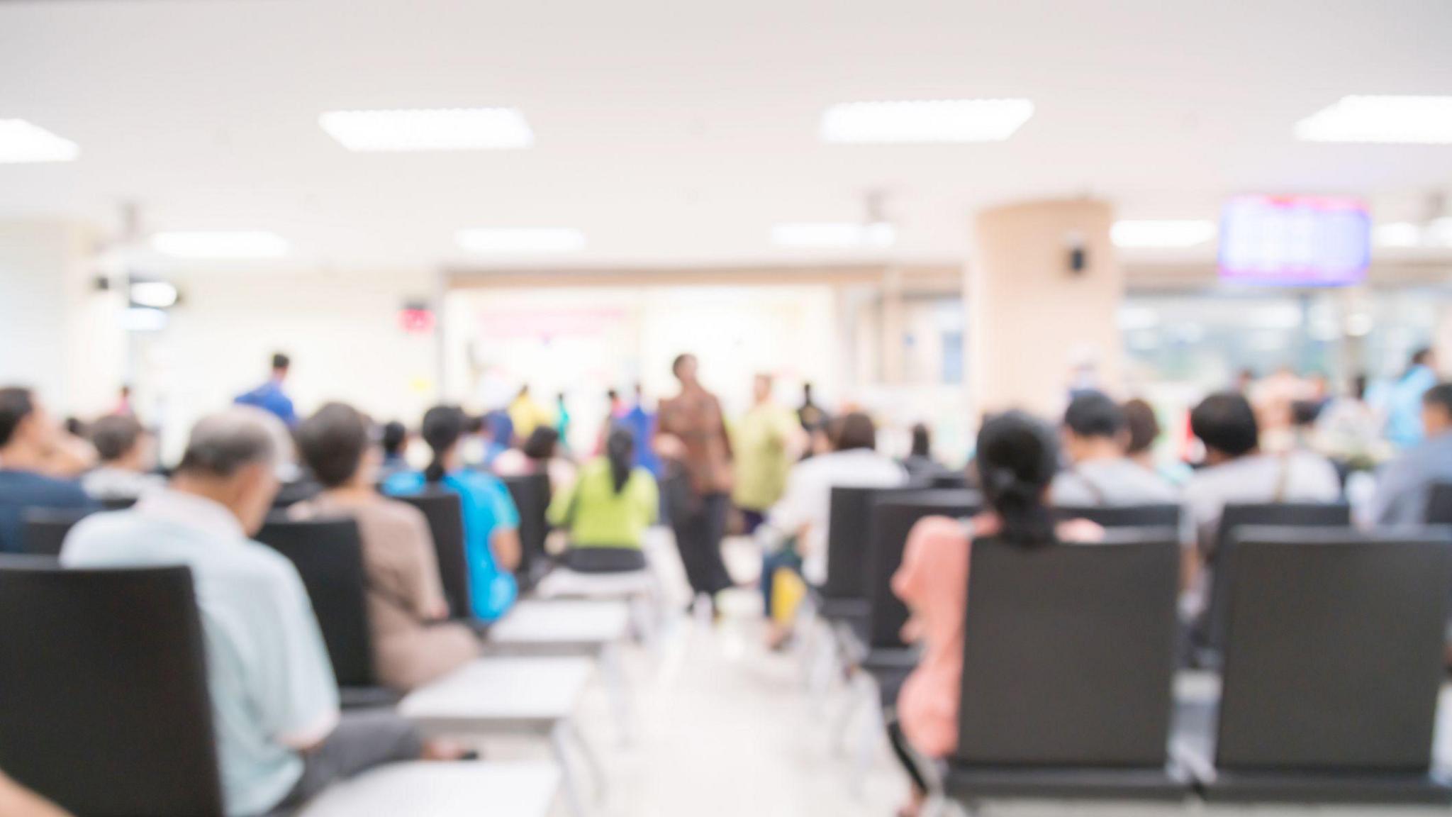 A blurred image of a busy hospital waiting room. There are many people sitting on chairs. A TV can be seen in the distance. 