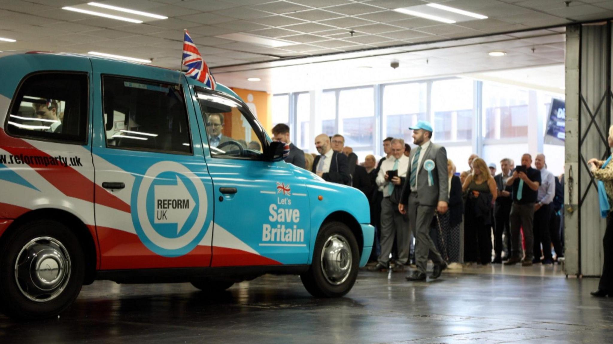 An image of a black cab covered in Reform UK branding at the Reform UK conference in Birmingham. The cab has the Reform turquoise logo, a union jack and the message "let's save Britain". There are a queue of delegates behind, including a man in a turquoise baseball cap and rosette.