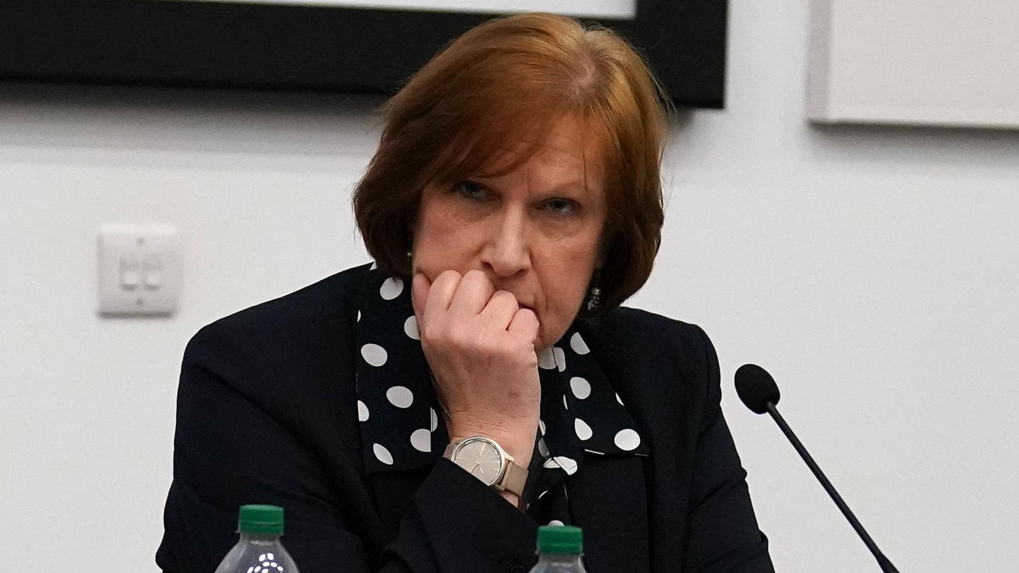 A woman, former NHS executive Jane Grant, is sat behind a desk holding her clenched hand against her face. She is sat in front of a microphone and is wearing a black jacket and polka dot shirt under that. 