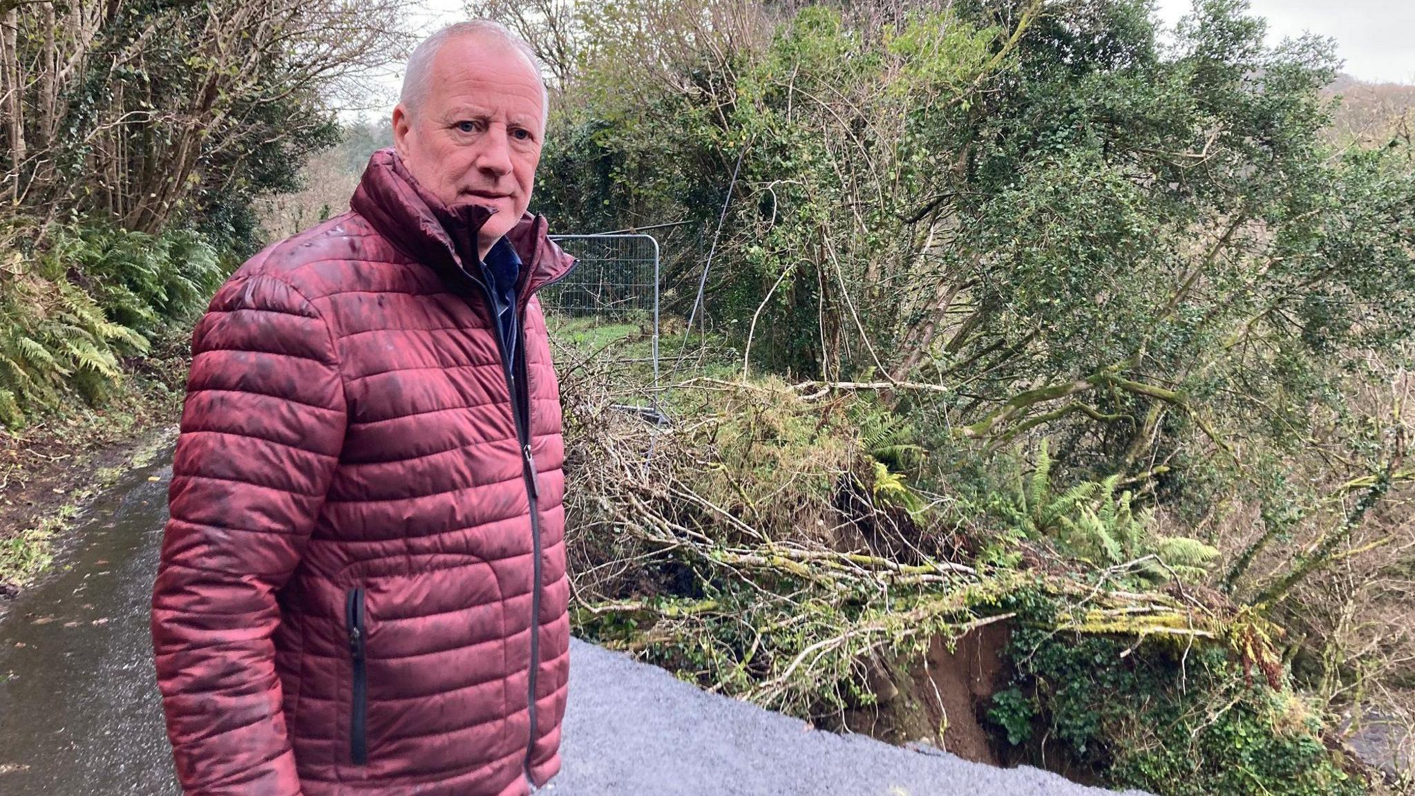 Frankie McBride standing beside a landslide on Glenshesk Road,  He has short, grey hair and is wearing a red padded jacket.   Part of the road behind him has fallen away. 