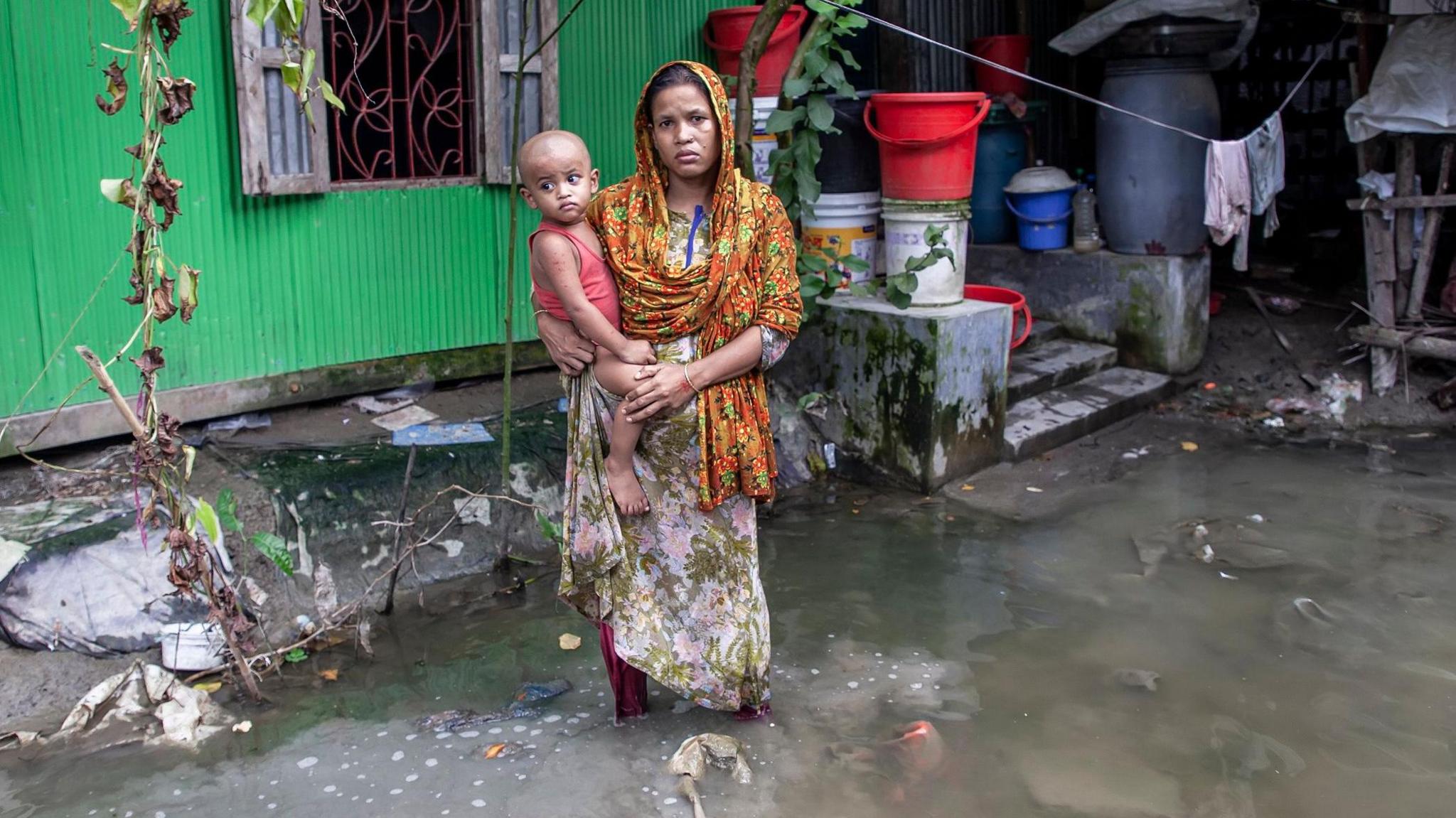 A woman holds a small child in her arms in front of her flooded house in a village in Bangladesh