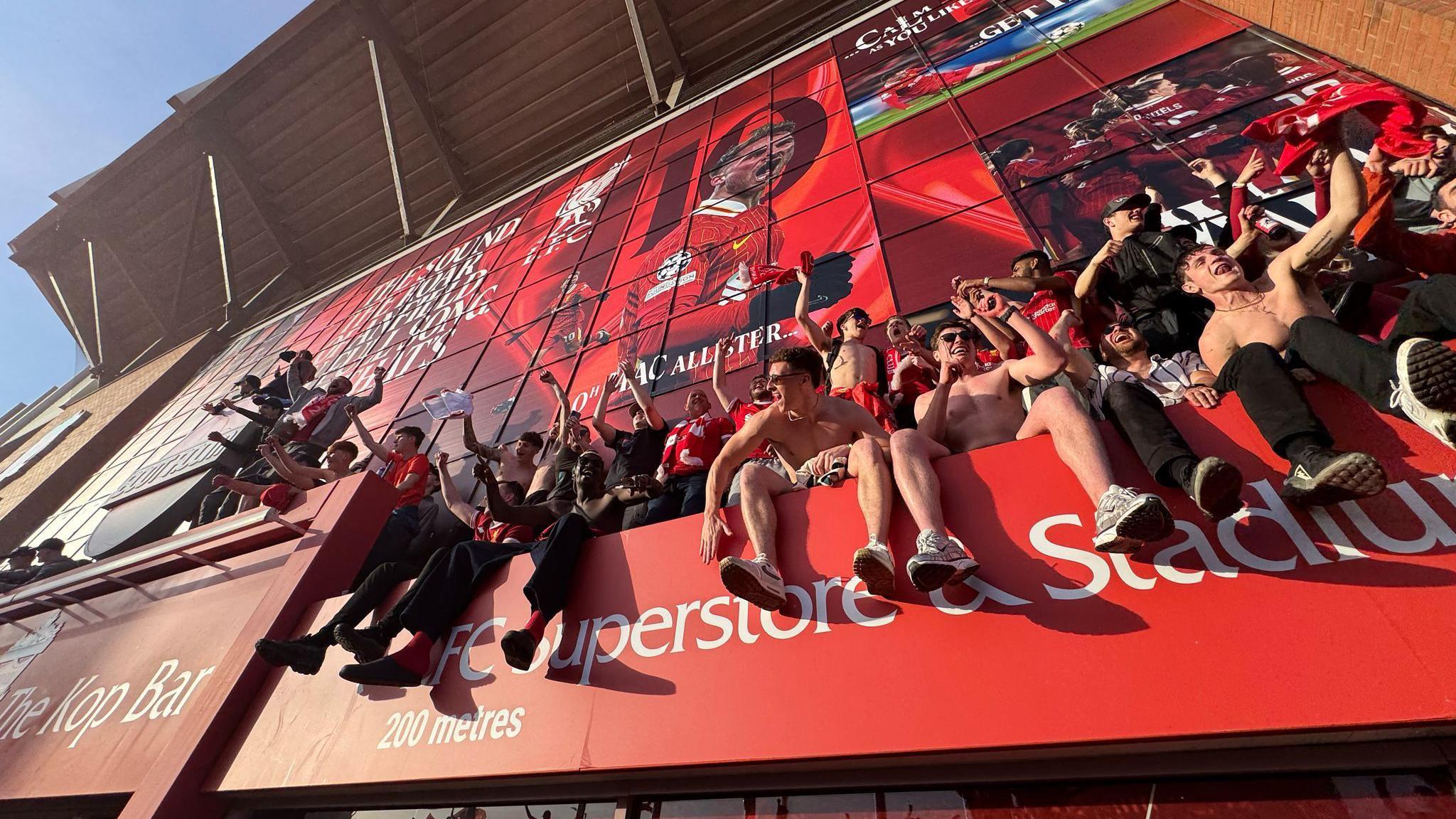 Fans, many with arms raised in the air, sitting on the sign for the Liverpool superstore at Anfield stadium as the match came to an end.