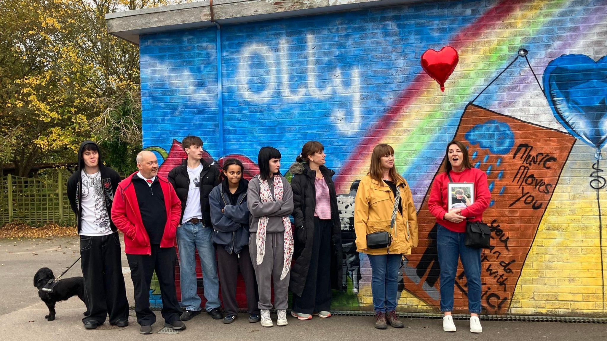 A line of eight people stood in front of a colourful wall. The wall has the word 'Olly' in big white writing on a blue background. There is also a rainbow and blue heart shaped balloon on the wall.