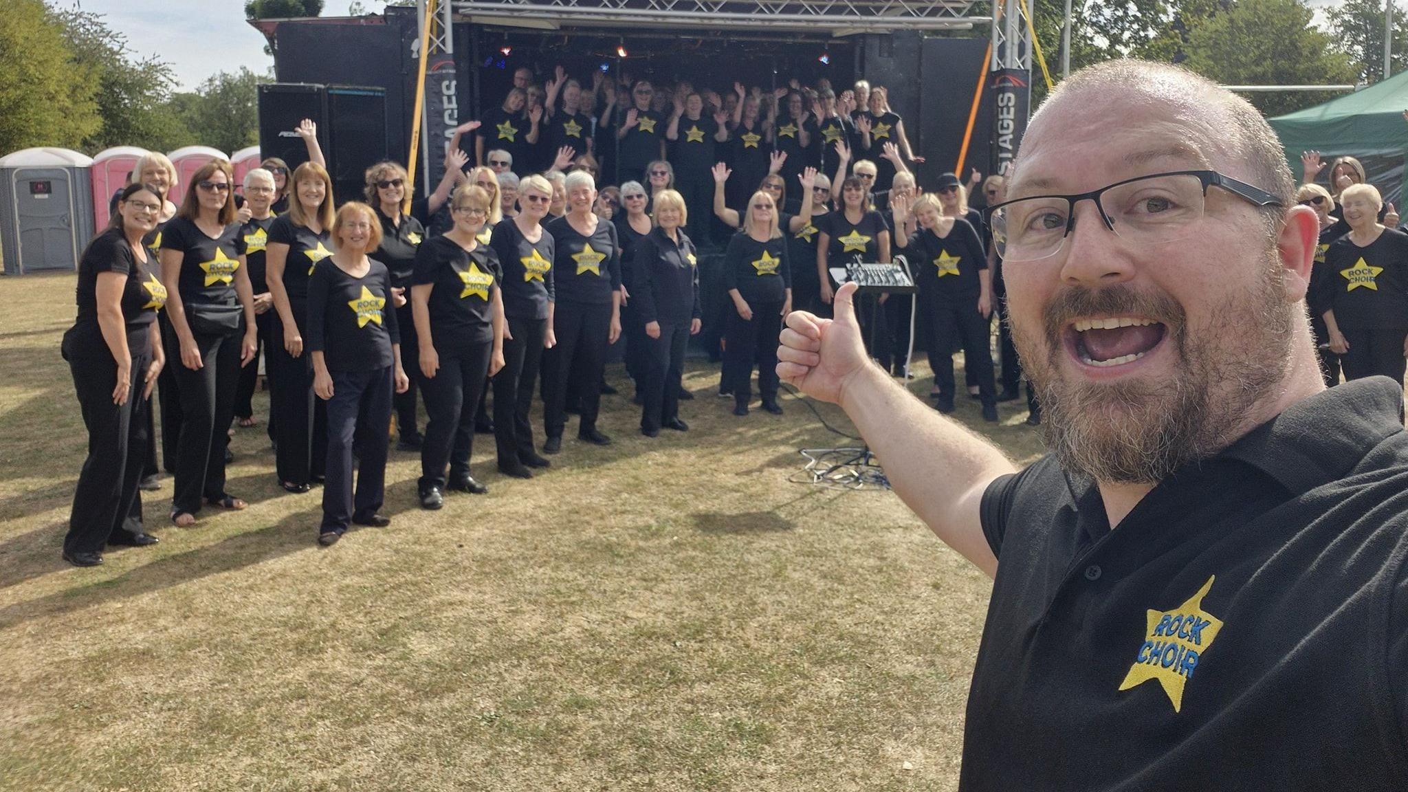 A man in a black polo taking a selfie with lots of choir members on a stage and off stage in the background. They are at a festival.