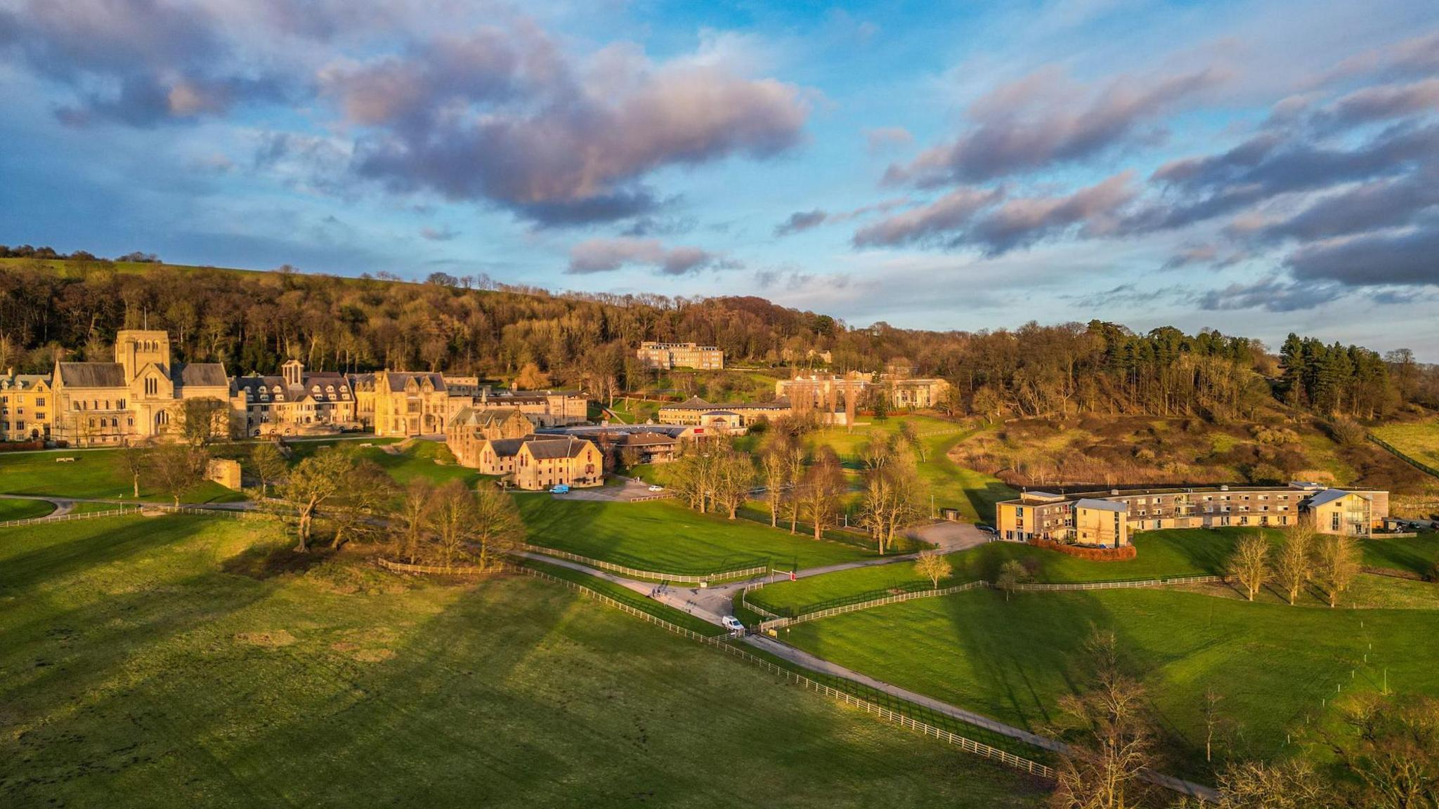 View of buildings spread across grounds, with trees and playing fields. Behind the buildings is a wooded area. Blue skies and clouds.