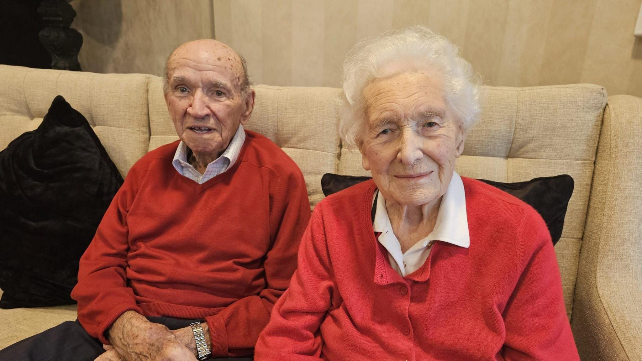 Alan and Trudy are sitting next to each other on a cream sofa, they are smiling at the camera and are both wearing red jumpers over white collared shirts. 