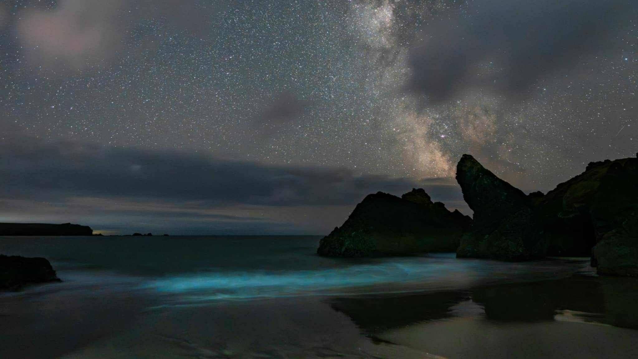 A picture of the view across Cornwall. In the background the sky full of stars can be seen with a body of water and shadow of cliffs surrounding it. At the bottom of the image a strong blue glow can be seen.