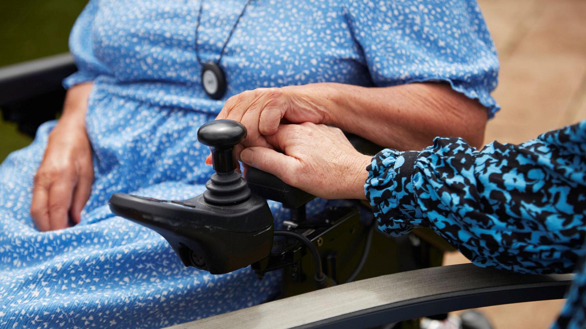 A picture of a women in a wheelchair holding hands with another women. Both are wearing blue patterned dresses and only their hands and part of a chair and a wheelchair can be seen. 