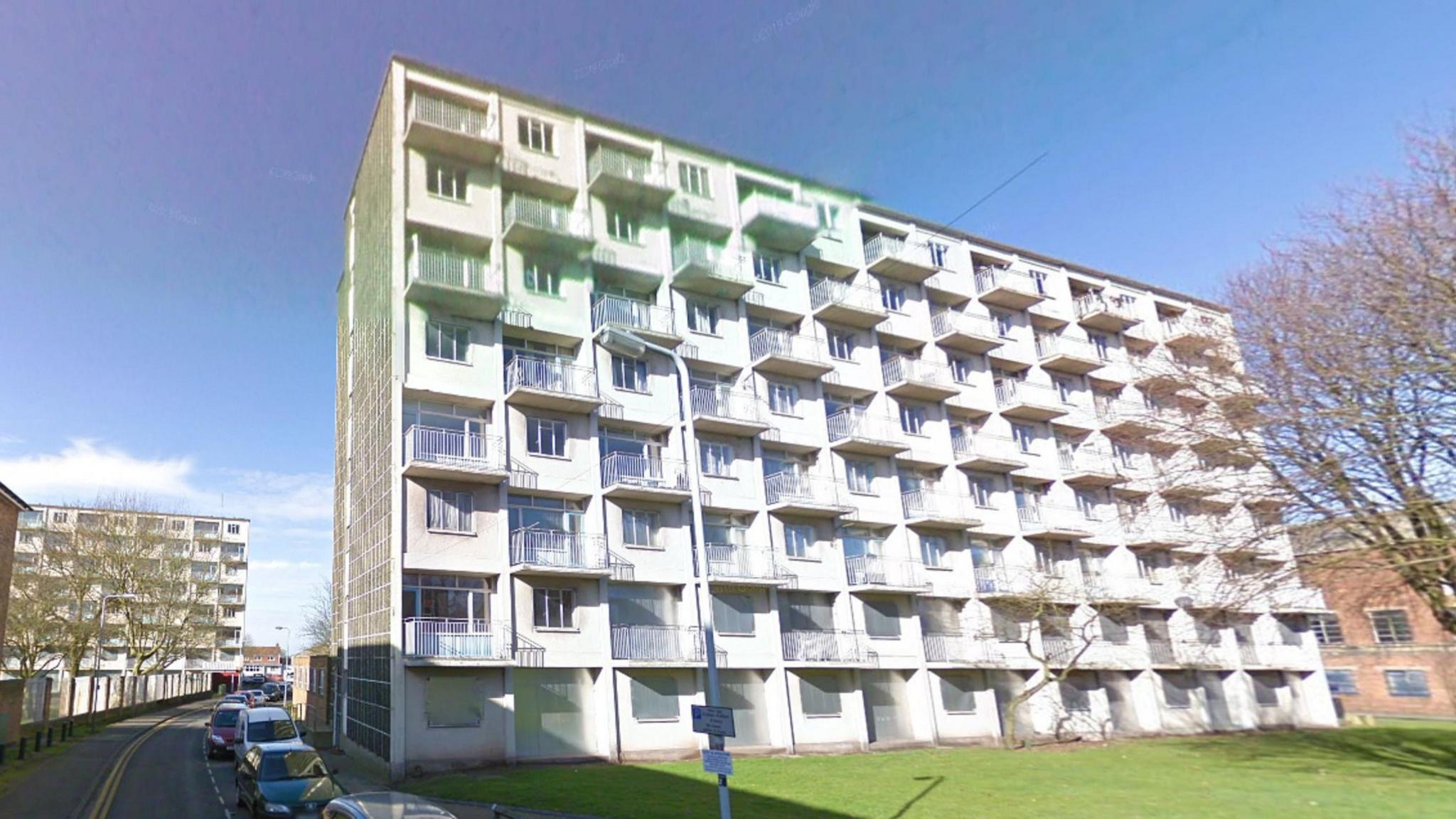 A large white block of flats sitting on a green space, surrounded by houses and other blocks of flats. The sky is blue, and cars are parked on the road which runs to the left of the block.