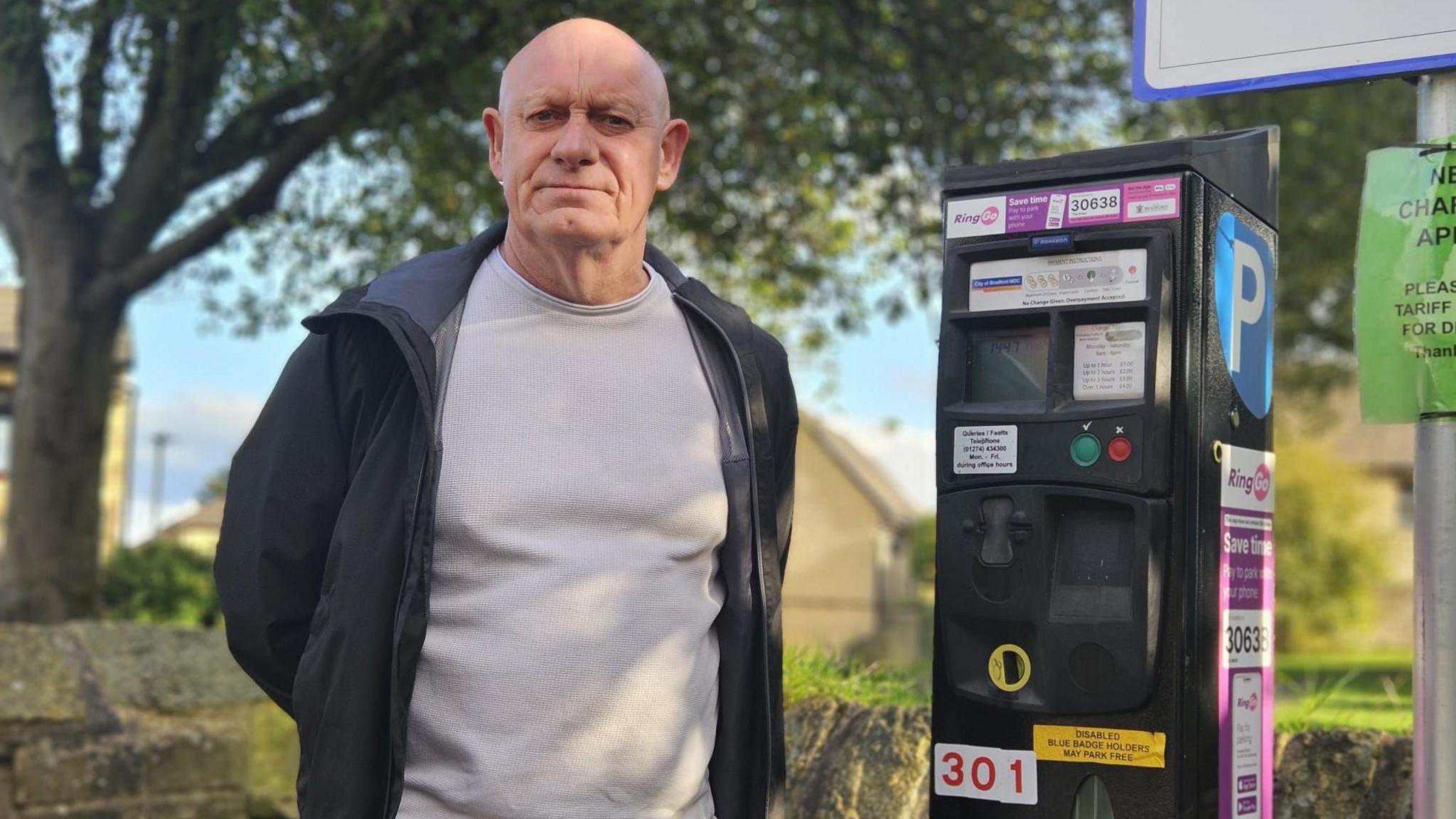 A man standing in front of a parking ticket machine, looking serious and with his hands behind his back.