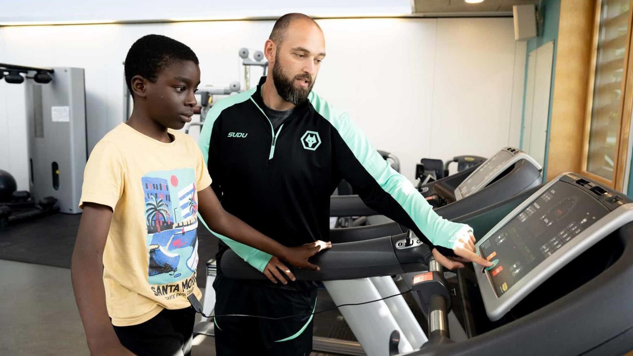 A boy takes part in a Yo! Active youth gym session. He is on a treadmill with a trainer learning how to work the machine. The trainer is pointing to the buttons and the boy is listening. There is gym equipment all around them.