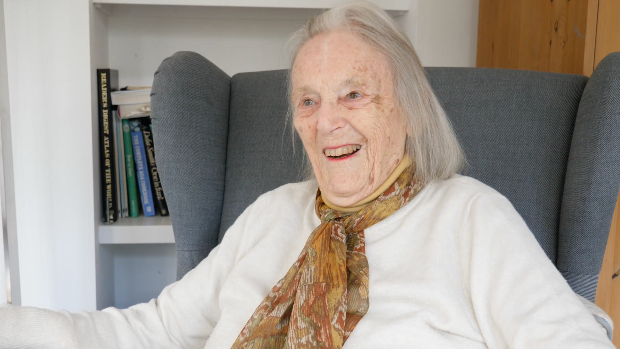 Rosemary Hanbury sat on a grey armchair wearing a white jumper with a yellow, green and red scarf. She is smiling and looking to the left. There is a book shelf with books on the left behind the chair.