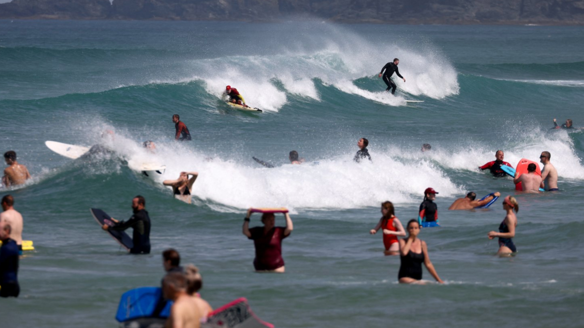 A number of surfers and swimmers in the sea, some are holding bodyboards and some are surfing.