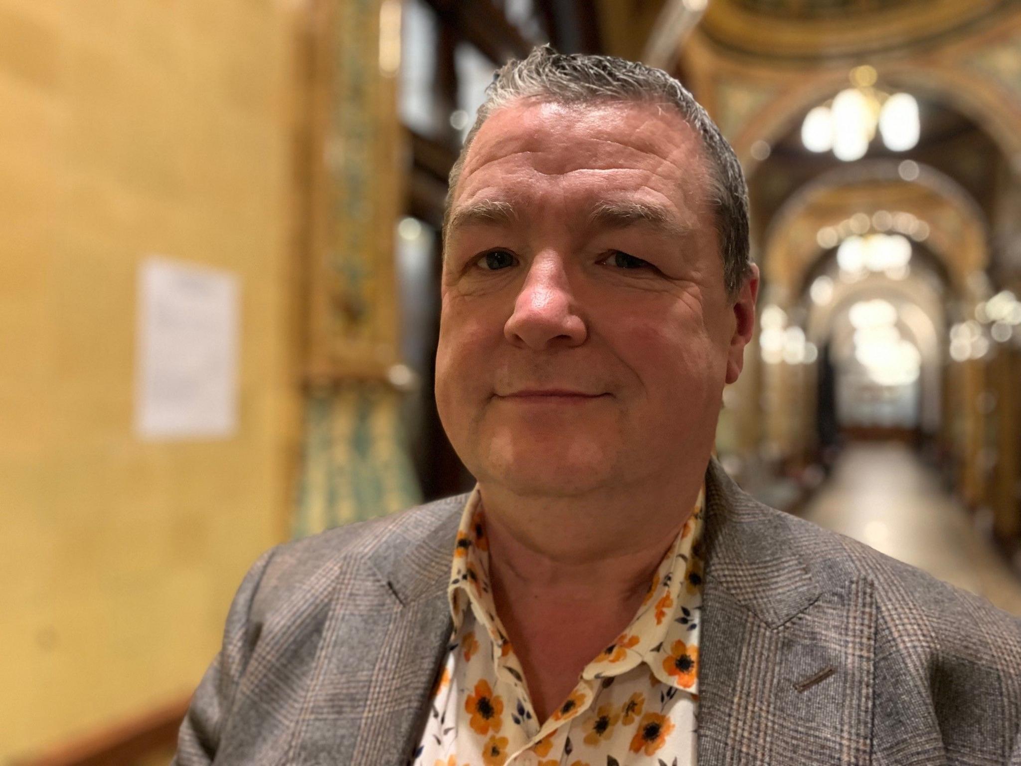 Cllr Richard Bell - man with short grey hair standing in a brightly lit corridor with arched ceiling, wearing a grey jacket and white shirt with yellow floral pattern